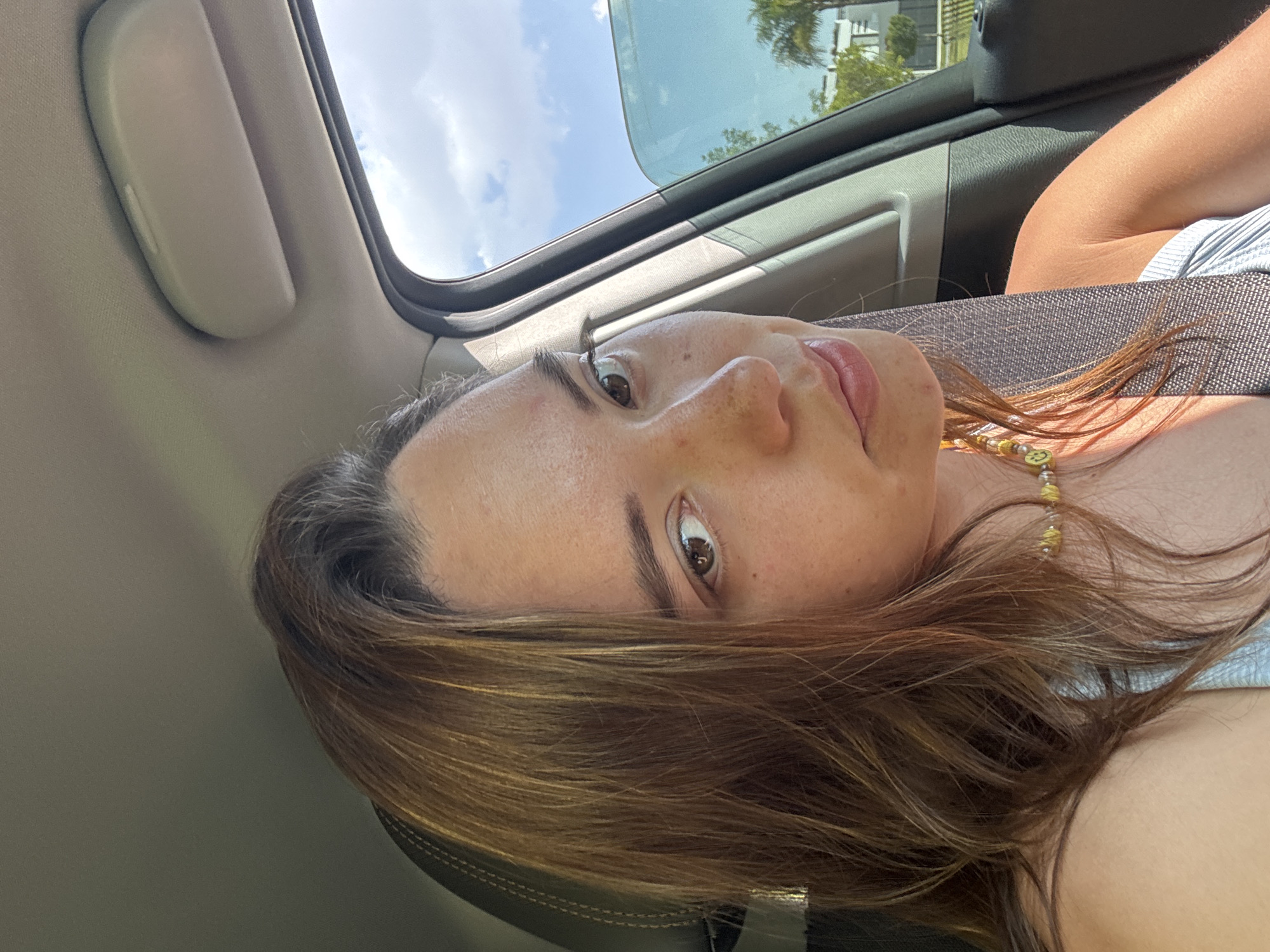 A woman is in a car. The car has a sunroof. The woman is wearing a seatbelt. Outside the car are trees and a building. There is no information in the image to determine if the location is San Bernardino Tlaxcalancingo, Mexico.