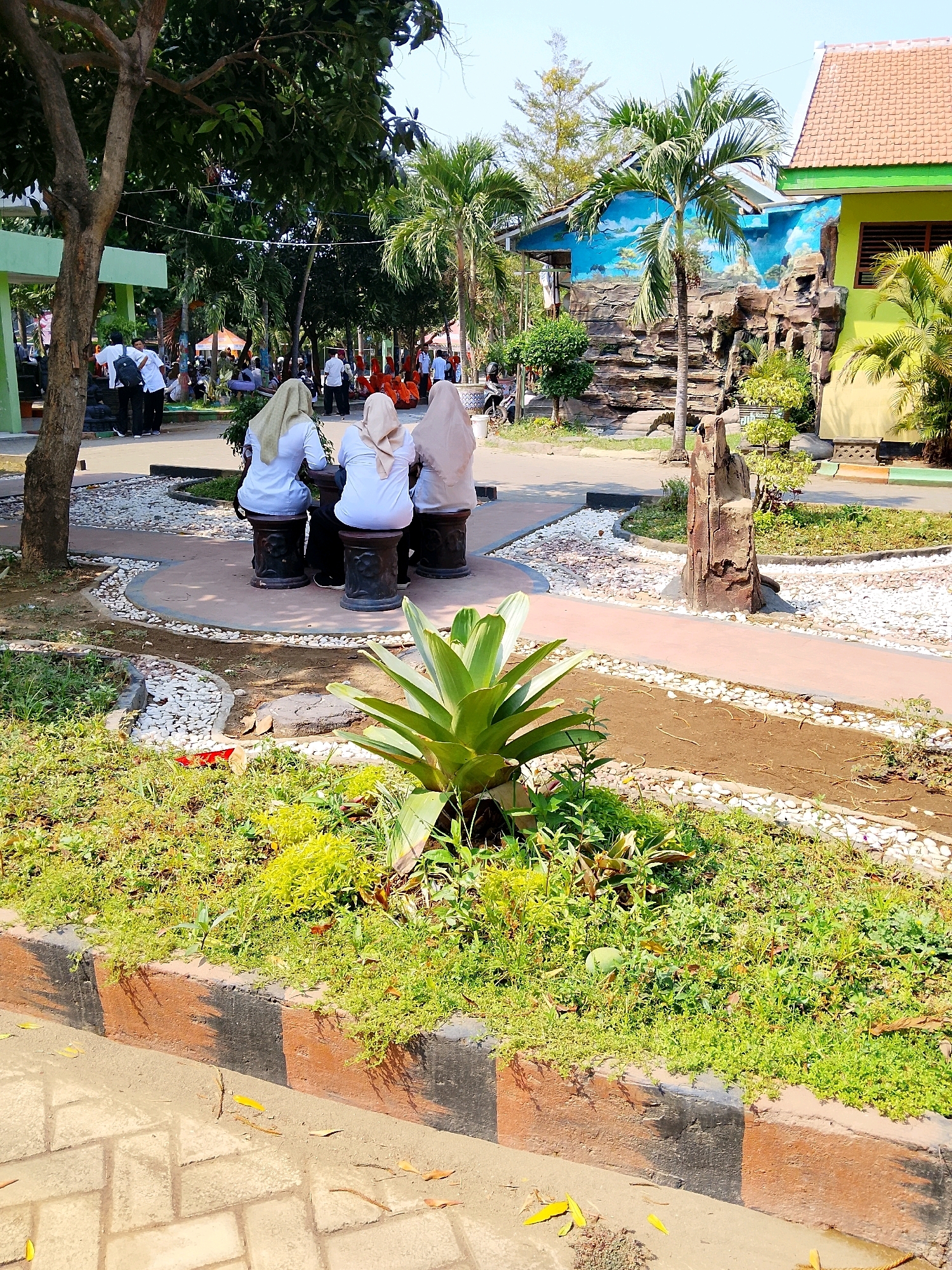 The image depicts an outdoor scene on a sunny day, likely at a school or public institution in Nganjuk, Indonesia, given the presence of students in uniforms and the tropical vegetation. In the foreground, a paved walkway made of light-colored bricks is visible, with fallen yellow leaves scattered on it. A low, painted curb, with alternating brown and black stripes, separates the walkway from a grassy area. This grassy area is densely populated with small, green plants and a larger, spiky plant with green leaves in the center.
Further into the scene, a circular paved area features three young women wearing white tops and light-colored hijabs, seated on dark, cylindrical stools. They are facing away from the camera, engaged in conversation or looking at something together. A low, white stone border surrounds this circular seating area, with a path of red gravel curving around it.
Beyond this seating area, more foliage is present, including palm trees. In the background, a building with a blue mural depicting a landscape with mountains and clouds is visible. This building has a yellowish-green facade. Other people, appearing to be students, are seen in the middle ground, some standing and some seated in an open area with orange seating. The overall environment suggests a well-maintained outdoor space, possibly a campus courtyard or a public park with educational facilities.