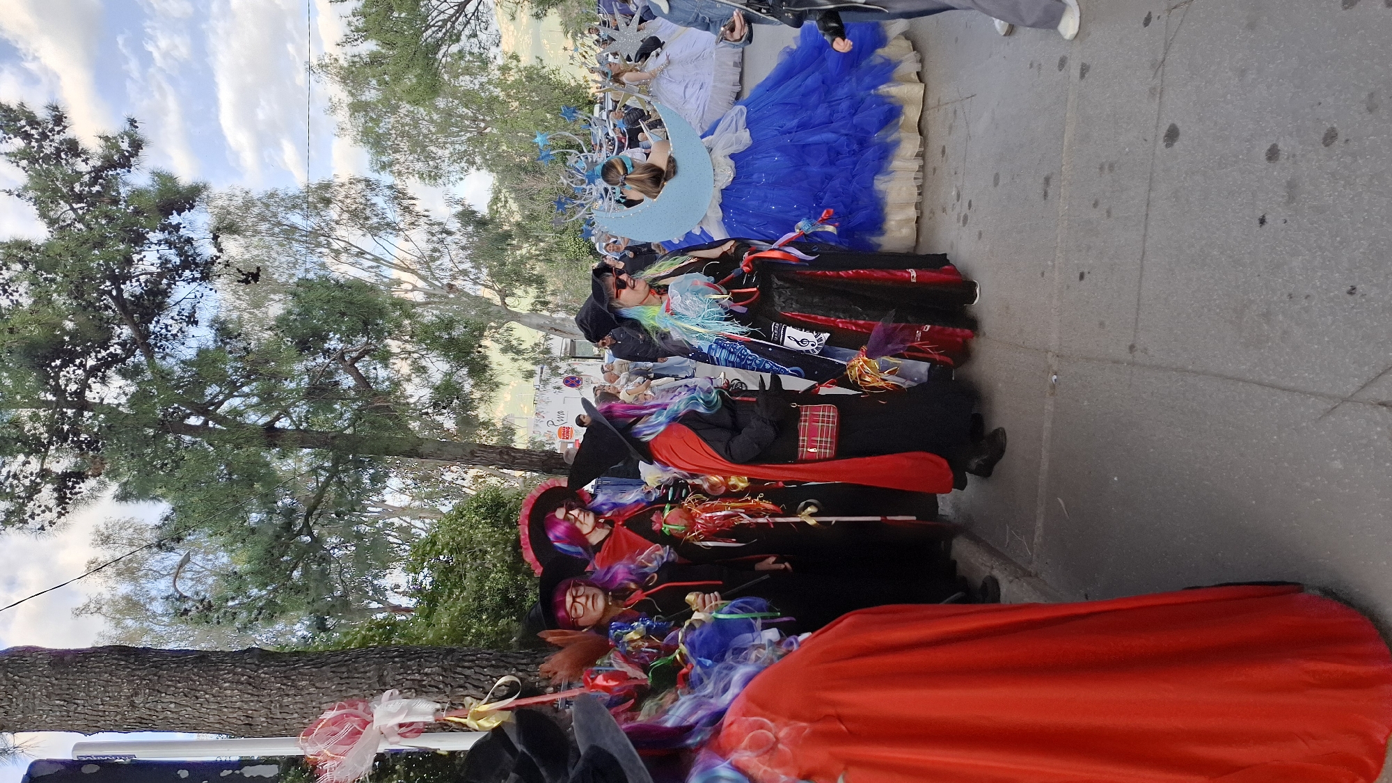 A vertical, medium shot depicts a parade or street performance in Budva, Montenegro. A group of individuals dressed in elaborate costumes are the primary focus, lined up along the pavement. The costumes are primarily black and red, with flourishes of blue, silver, and rainbow colors in the wigs and accessories. One person wears a blue gown, adorned with a large crescent moon and stars. A tall tree stands to the left, its branches and leaves partly obscuring the sky. The sky is visible in patches, with white clouds against a pale blue background. A paved road stretches into the distance, with pedestrians partially visible. The photograph is taken from a slightly elevated viewpoint.