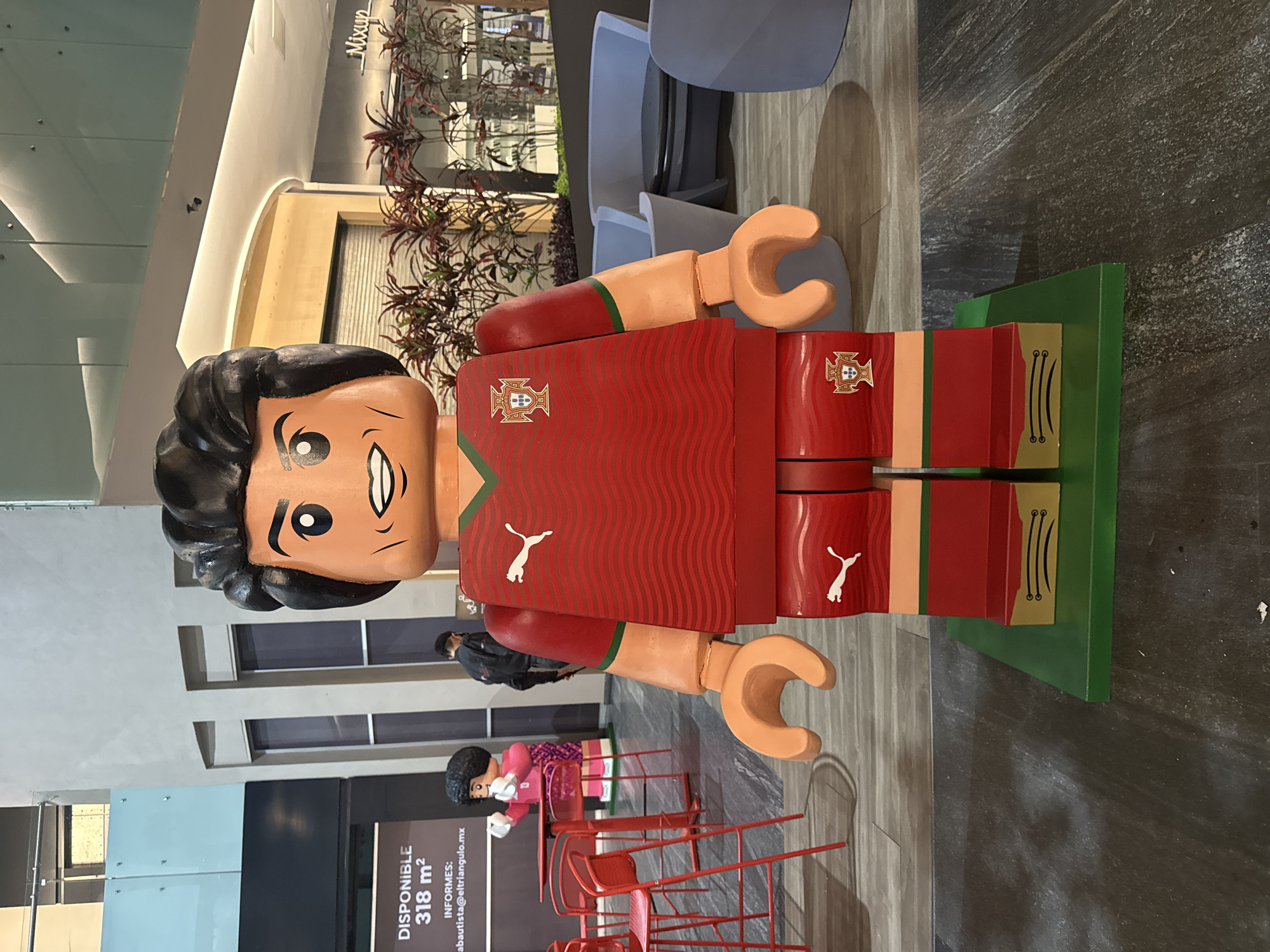 A large Lego figure, styled to represent Cristiano Ronaldo in a Portugal soccer jersey, stands on a green base in a brightly lit commercial space. The figure is predominantly red with the Portuguese coat of arms and a Puma logo visible. The location appears to be a shopping mall or commercial area, given the presence of furniture, plants, and storefronts in the background. One sign that is visible advertises that there are 318 square meters of space available for rent, including an email address for inquiries. People are walking by the figure. The scene is in Puebla, Mexico, judging from the advertisement.