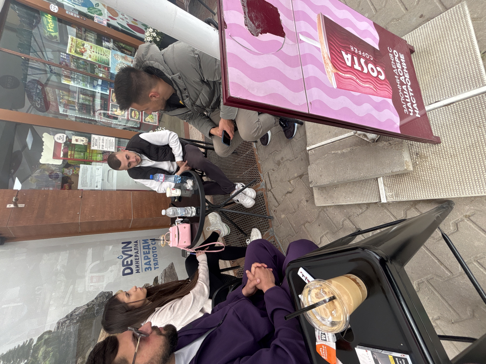 The image shows a group of people sitting at tables outside a Costa Coffee location in what appears to be Plovdiv, Bulgaria. The photo is taken from an elevated angle, looking down on the scene.
Several people are seated around small tables. One table is branded with Costa Coffee advertisements. There are various drinks, including iced coffee in plastic cups, and water bottles on the tables. Several individuals are looking at their phones, and some seem to be in conversation.
The setting is an outdoor seating area in front of a building with a glass facade. The facade features signs advertising "Fresh" produce and other goods. In the background, advertisements for "Devin" mineral water are visible. The environment suggests an urban setting, likely in the center of Plovdiv.