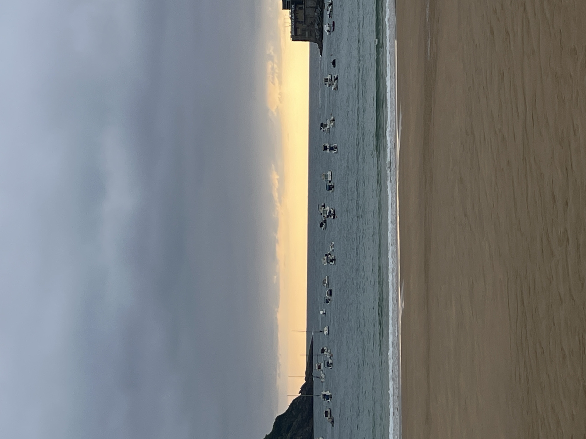 The image shows a panoramic view of La Concha Bay in San Sebastián, Spain, at dusk. The sky is overcast with dark clouds, but a band of soft orange and yellow light is visible near the horizon, indicating the setting sun. Numerous small boats are anchored in the calm waters of the bay, some with masts visible. On the left side of the image, a lush green hillside slopes down towards the water, with a few buildings nestled at its base. On the right side, a stone structure, possibly a fortification or an old building, juts out into the sea. The foreground is dominated by a sandy beach with gentle waves lapping at the shore. The scene conveys a sense of tranquility and natural beauty, typical of this popular tourist destination.