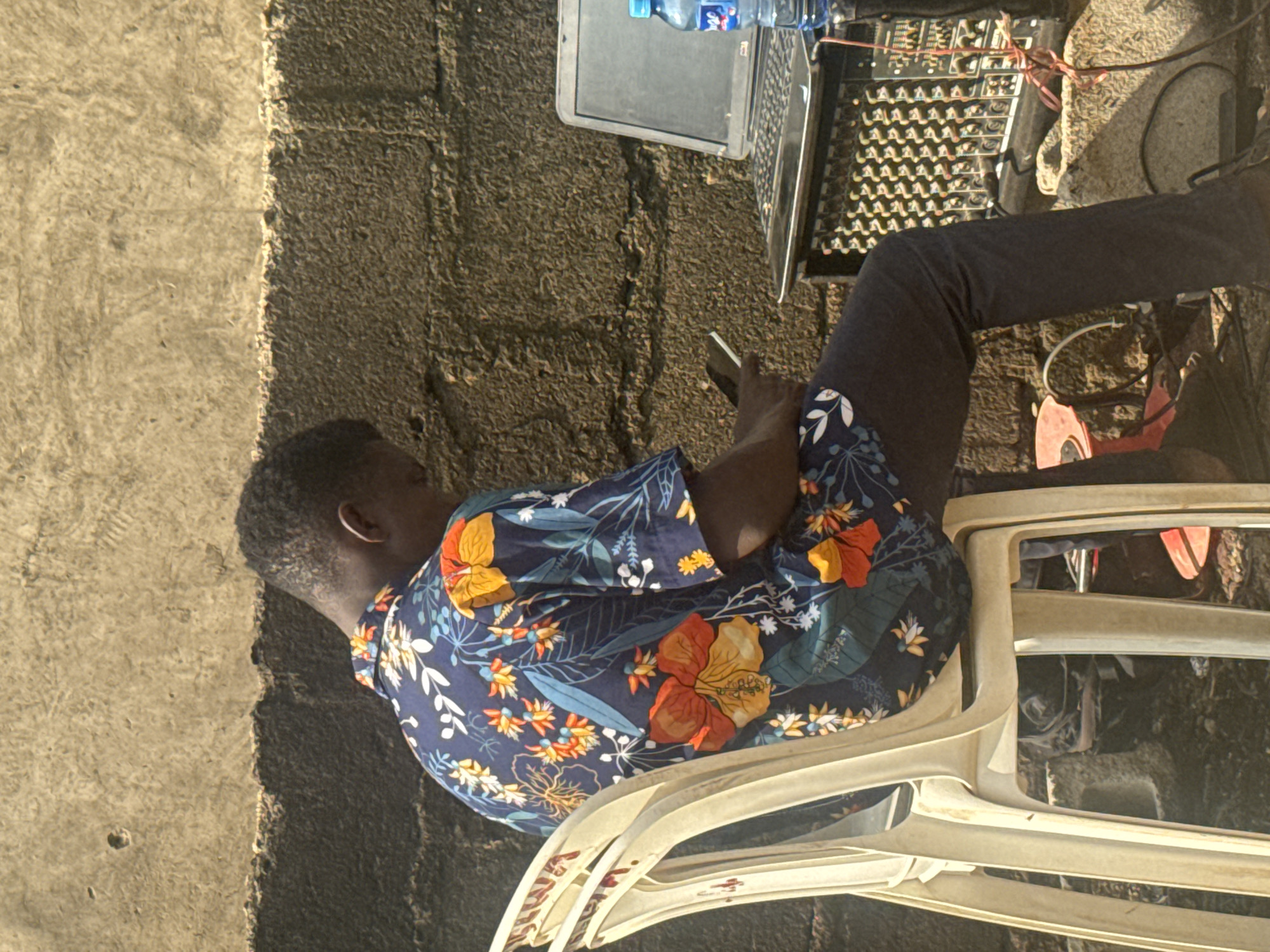 In Jawu, Nigeria, a young man sits on a white plastic chair, facing away from the camera, engrossed in his smartphone. He is wearing a dark blue short-sleeved shirt adorned with a vibrant floral pattern of orange, yellow, and white flowers. His dark pants contrast with the bright shirt. Behind him, a textured, weathered stone wall serves as the backdrop, illuminated by natural sunlight that casts shadows. To his right, a laptop rests on top of a black audio mixing board, with a blue bottle of water placed nearby. The scene suggests an outdoor setup, possibly for a public event or a workspace.