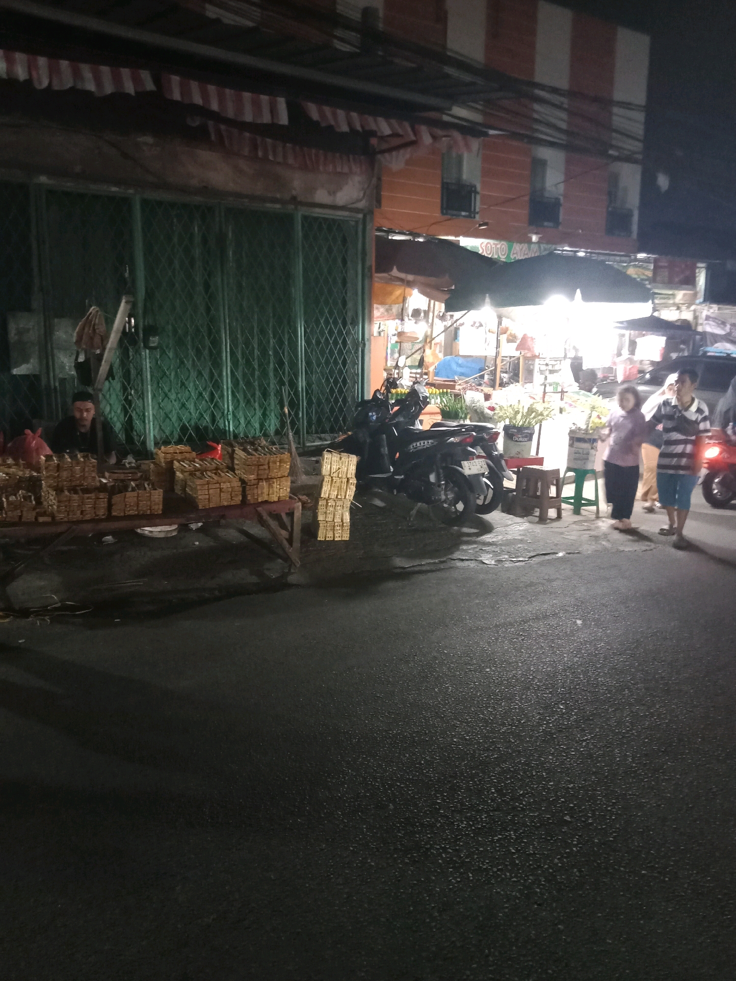 An outdoor night market scene in South Tangerang, Indonesia, is depicted, focusing on a vendor stall in the foreground. A man wearing a black shirt is seated behind a table laden with stacks of what appear to be pre-packaged food items, possibly snacks or desserts, wrapped in brown paper. A red plastic bag is visible to his left. The stall has a green metal gate in the background. To the right of the stall, several motorcycles are parked. Further to the right, three people are standing near other stalls, illuminated by overhead lights. One person, a woman in a purple shirt and dark pants, stands with her hands clasped. Beside her, a person in a striped shirt and blue shorts faces the viewer, and another person is partially visible to their right. The ground is paved and wet, reflecting the ambient light. The market appears to be located in a commercial area with buildings and awnings visible in the background.