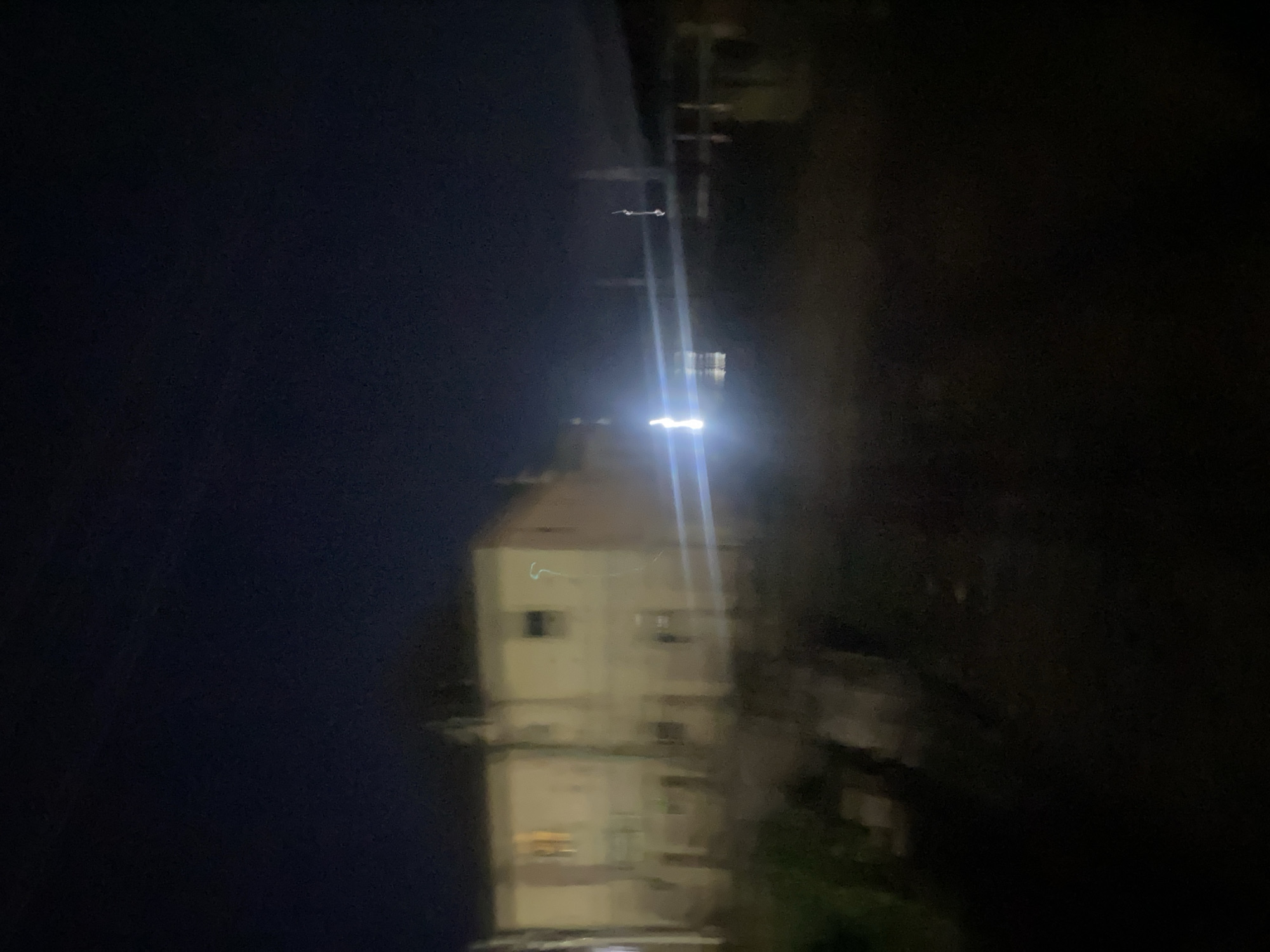 The image, taken at night, depicts a blurry, overexposed view of buildings in Jos, Nigeria. The structures appear to be two or three stories high and are illuminated by what seems to be artificial lighting, creating strong lens flares that obscure details. The foreground is dark and out of focus, suggesting a natural or undeveloped landscape. The overall impression is one of low visibility and atmospheric conditions that have degraded the photographic quality.