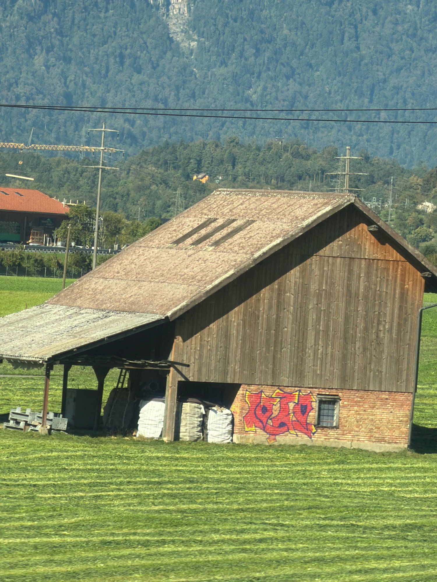 A wooden barn with a tin roof stands in a green field in Steinen, Switzerland. The barn has a brick base and a large open section on the left. Red and yellow graffiti is spray-painted on the brickwork. There are several large white bales of hay or straw stacked near the open section of the barn. In the background, a dense forest covers a mountain slope. Power lines and utility poles are visible in the middle ground. The field in front of the barn is a vibrant green, with visible rows of cut grass, suggesting recent mowing. The sky is a clear, pale blue.