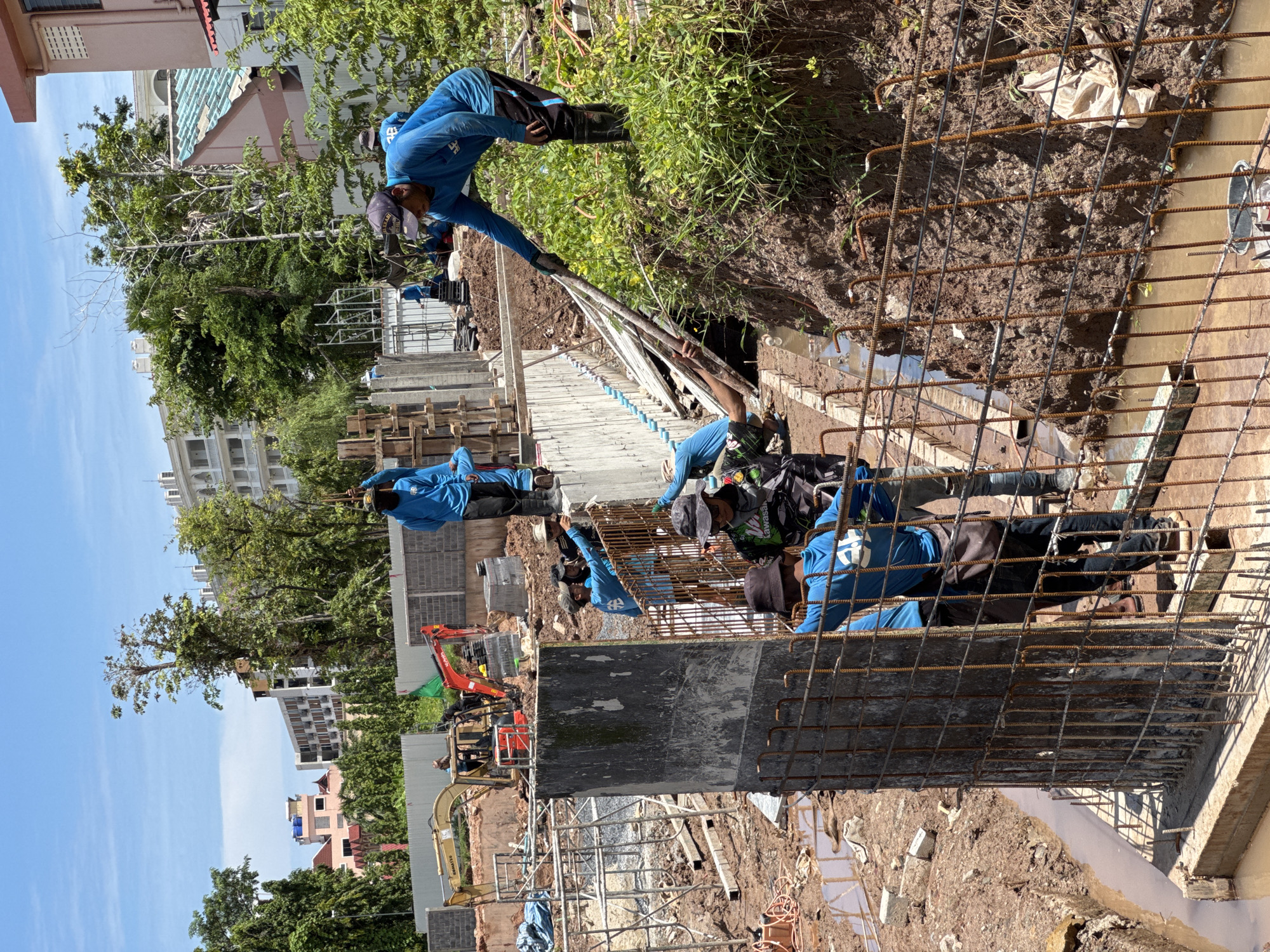 The image depicts a construction site in Patong, Thailand, on a sunny day. Several workers, dressed in blue long-sleeved shirts and dark pants or shorts, are engaged in various construction activities. The foreground shows a concrete structure with rebar reinforcement, surrounded by a rusty metal fence. Muddy ground and standing water indicate recent rain or ongoing work. In the background, construction equipment including yellow and red excavators are visible, along with unfinished building structures and lush green trees. Several multi-story buildings, some appearing modern and white, are situated behind the construction site, suggesting an urban or semi-urban environment. The sky is clear blue with some clouds. One worker in the foreground appears to be directing another, while others are involved in moving materials and working on the concrete structure. The overall scene conveys active construction work in a tropical setting.