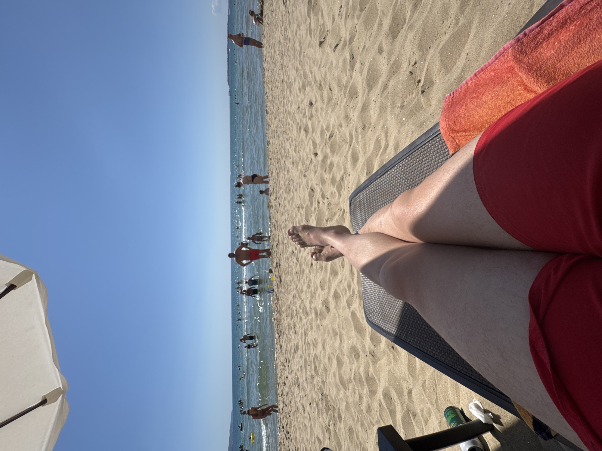 A first-person perspective shot captures an individual relaxing on a beach in Nesebar, Bulgaria. The foreground is dominated by the legs of the person, clad in red swim trunks, resting on a lounge chair. A portion of a beach umbrella is visible in the upper left corner. The midground shows a sandy beach populated by various beachgoers, some wading in the sea, others standing near the shore, and a child playing with sand. The background features a calm, blue sea under a clear sky, with a distant shoreline. The sun is shining brightly, casting shadows.