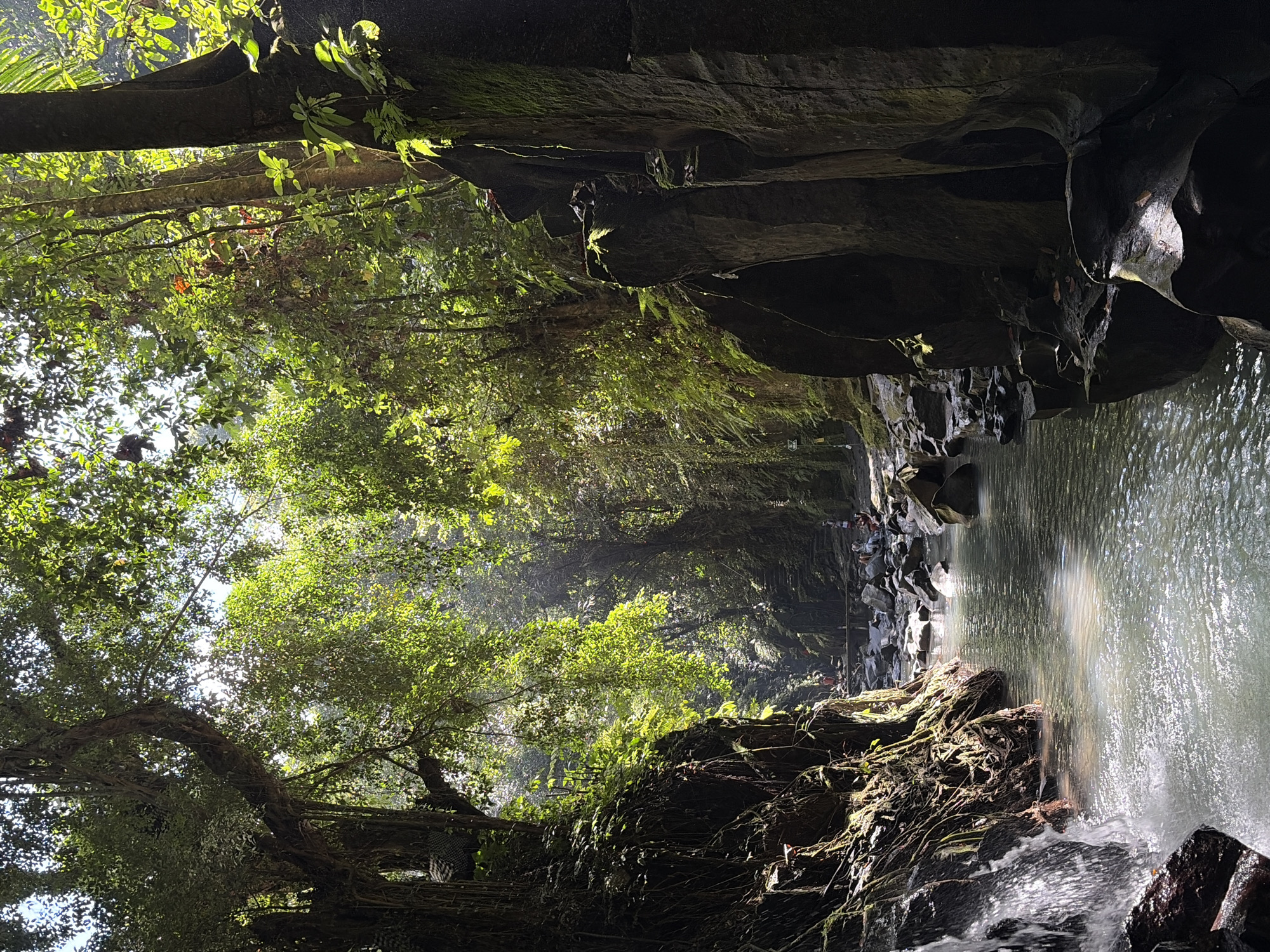 The image captures the Tukad Cepung Waterfall in Gianyar, Indonesia, a popular natural attraction. The scene depicts a narrow canyon with a stream flowing through it, flanked by towering, moss-covered rock walls. Lush green vegetation drapes from the canyon walls and overhead, with sunlight filtering through the dense canopy, creating dappled light effects on the water and rocks. A small waterfall cascades on the left side of the frame, adding to the serene atmosphere. In the distance, several people are visible, some standing near the water and others on what appears to be a wooden bridge or pathway, suggesting the area is frequented by tourists. The overall impression is one of natural beauty and tranquility, with the unique geological formations and the interplay of light and shadow contributing to the mystical ambiance of the location.