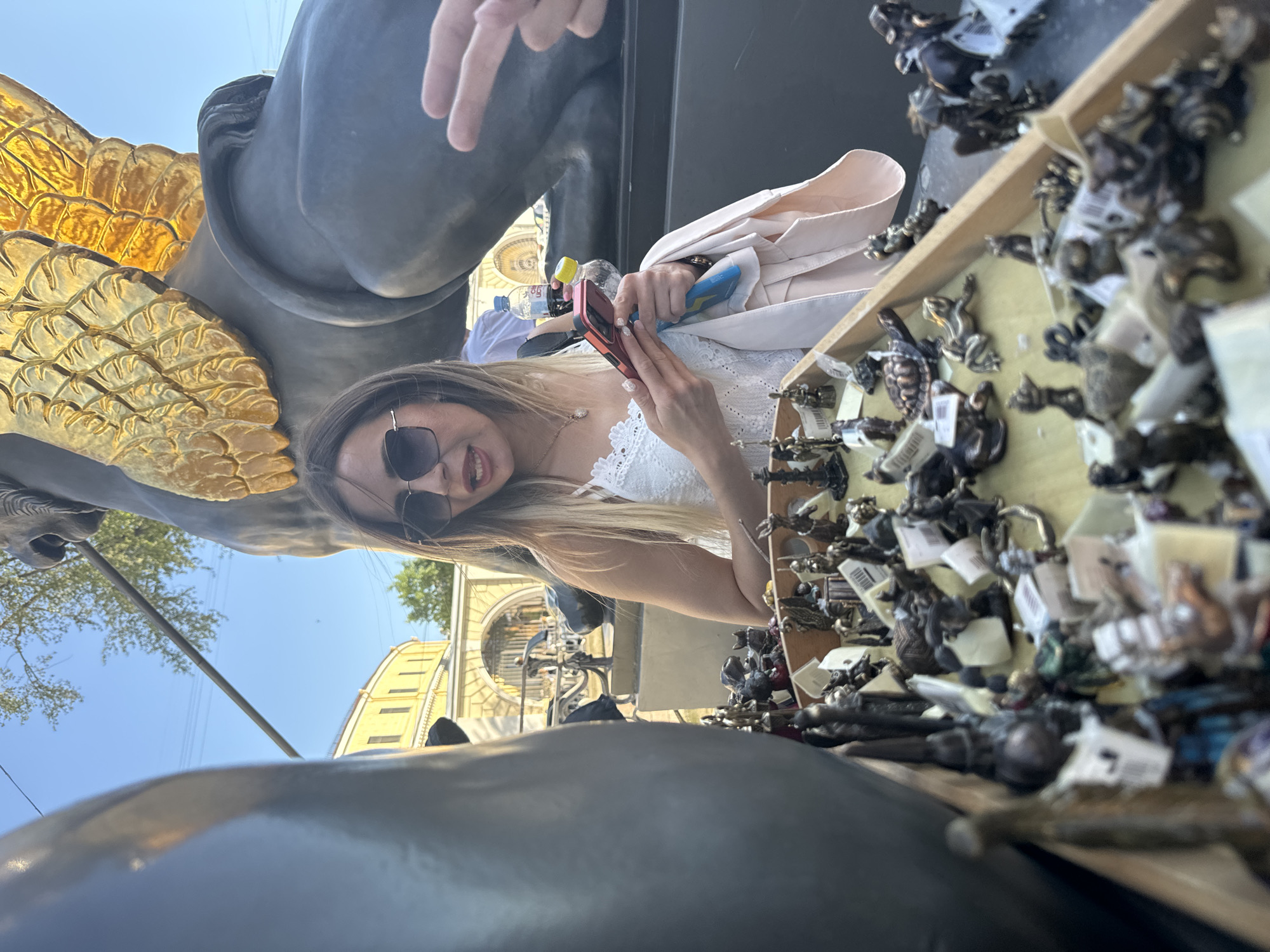 The image shows a young woman with blonde hair and sunglasses looking at the camera, smiling. She is wearing a white lace top and a light pink jacket. She is holding a pink smartphone in her hands and appears to be looking at it. In the background, a large statue of a winged creature with golden wings is visible, along with a stall displaying many small figurines of animals and mythical creatures.
The scene appears to be outdoors on a sunny day, as indicated by the bright sunlight and blue sky. The background suggests an urban setting with buildings that have classical architecture, consistent with the context of Saint Petersburg, Russia. The atmosphere seems casual and leisurely, possibly a tourist area or an outdoor market. The woman's expression suggests happiness and engagement with her surroundings or her phone. A bottle of water and a hand reaching into the frame are also visible. The small figurines are displayed in a wooden tray and many have price tags attached. The time of day appears to be late morning or afternoon given the sunlight.