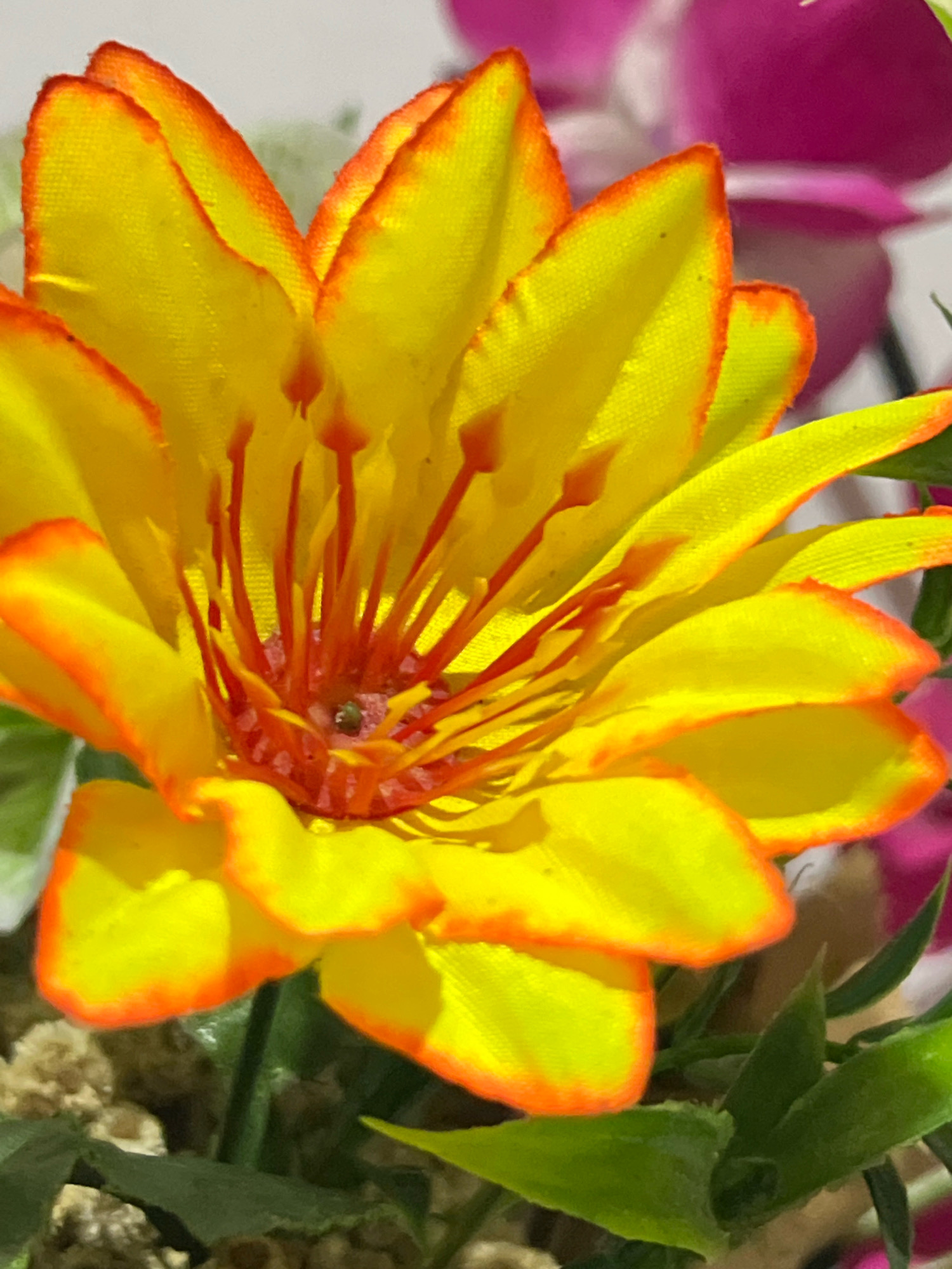 The image is a close-up shot of a bright yellow artificial flower with orange-red edges on its petals. The flower's center features numerous thin, red stamens. In the background, out-of-focus pink and white floral elements are visible, along with some green foliage and a textured brown surface that could be part of the flower arrangement or pot. The lighting suggests it is daytime, and the overall impression is of a vibrant, decorative floral display. There is no discernible information about its location in Denpasar, Indonesia, or any specific context beyond the visual details of the artificial flower.