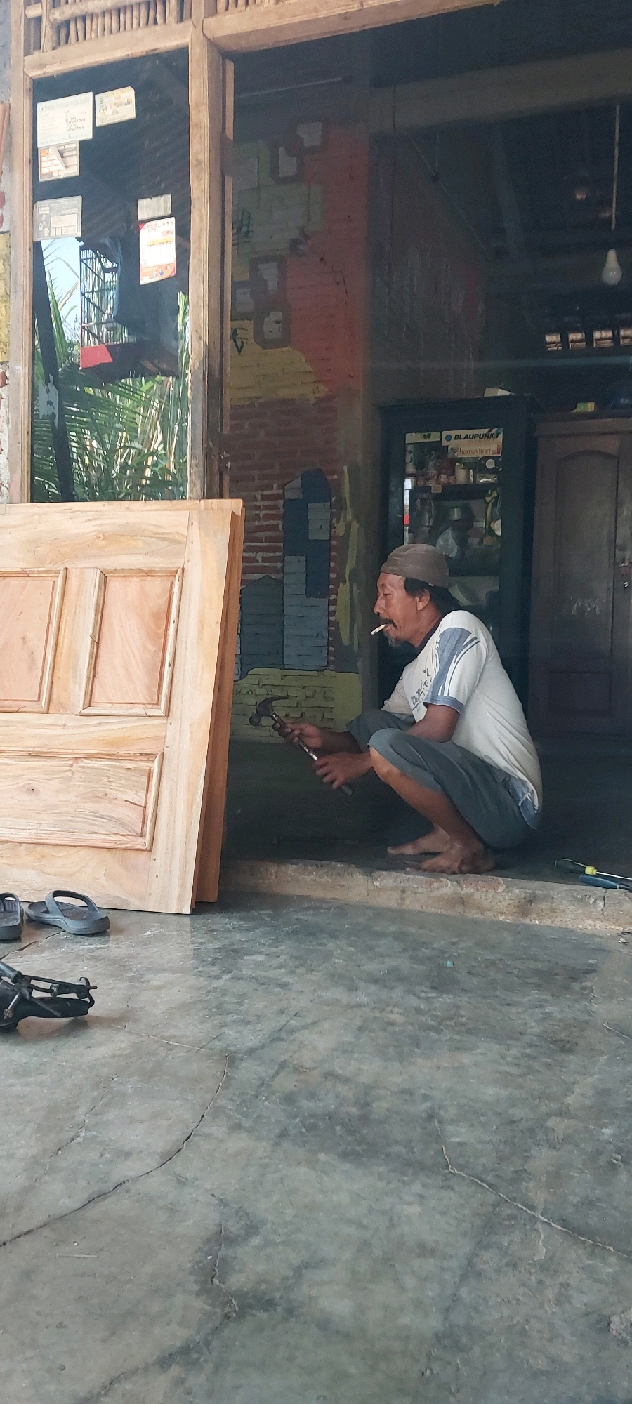 A man is squatting on a raised concrete step, holding a hammer in his right hand, preparing to work on a wooden door that is leaning against a wooden frame on his left. He is wearing a brown cap, a white t-shirt with blue stripes on the sleeves, and gray pants. A cigarette is visible in his mouth. To the left of the door are a pair of dark flip-flops and a black tool. The background shows a brick wall with colorful painted squares, and further back, a dark cabinet with a light-colored top. The floor is a cracked concrete surface. The scene appears to be outdoors, possibly in front of a workshop or building in Keling, Indonesia.