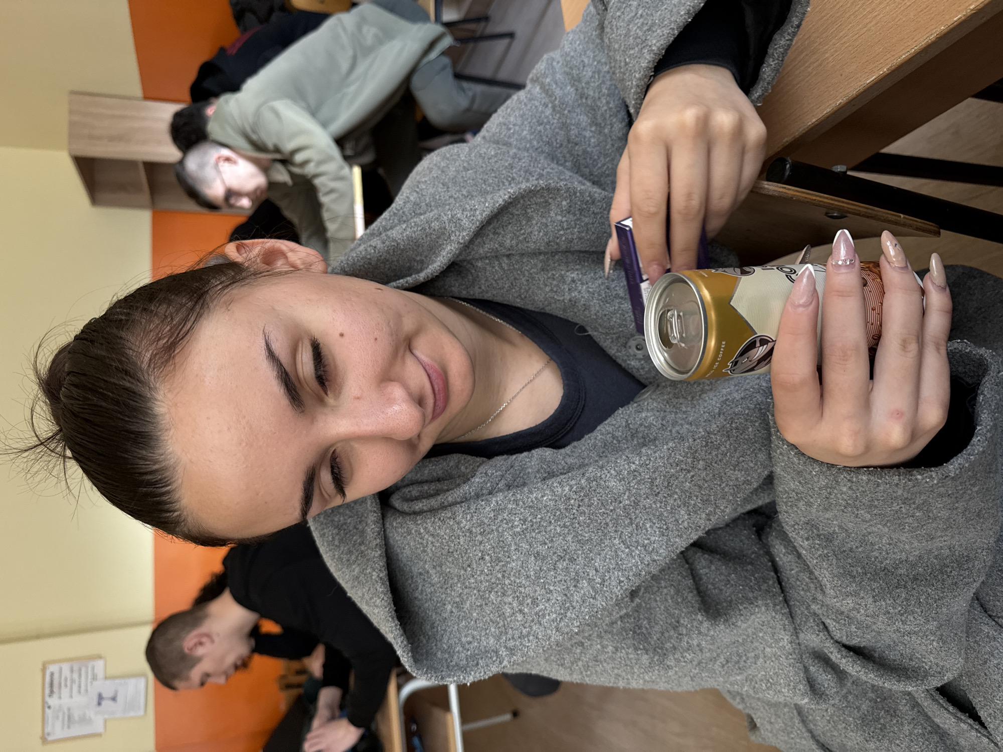 A woman is sitting at a wooden desk, holding a can of iced coffee in her right hand and a small, dark-colored rectangular package in her left hand. She is wearing a grey wool coat and a dark shirt with a silver necklace. She has dark hair pulled back in a bun and is looking down at the can. In the background, there are two other people. One is leaning over a desk, and the other is seated, looking at his hands. The room has orange and tan colored walls. There is also a white sheet of paper with black lettering and a symbol on the wall. The location is possibly a classroom or study area. There is no information to indicate it is in Sofia, Bulgaria.