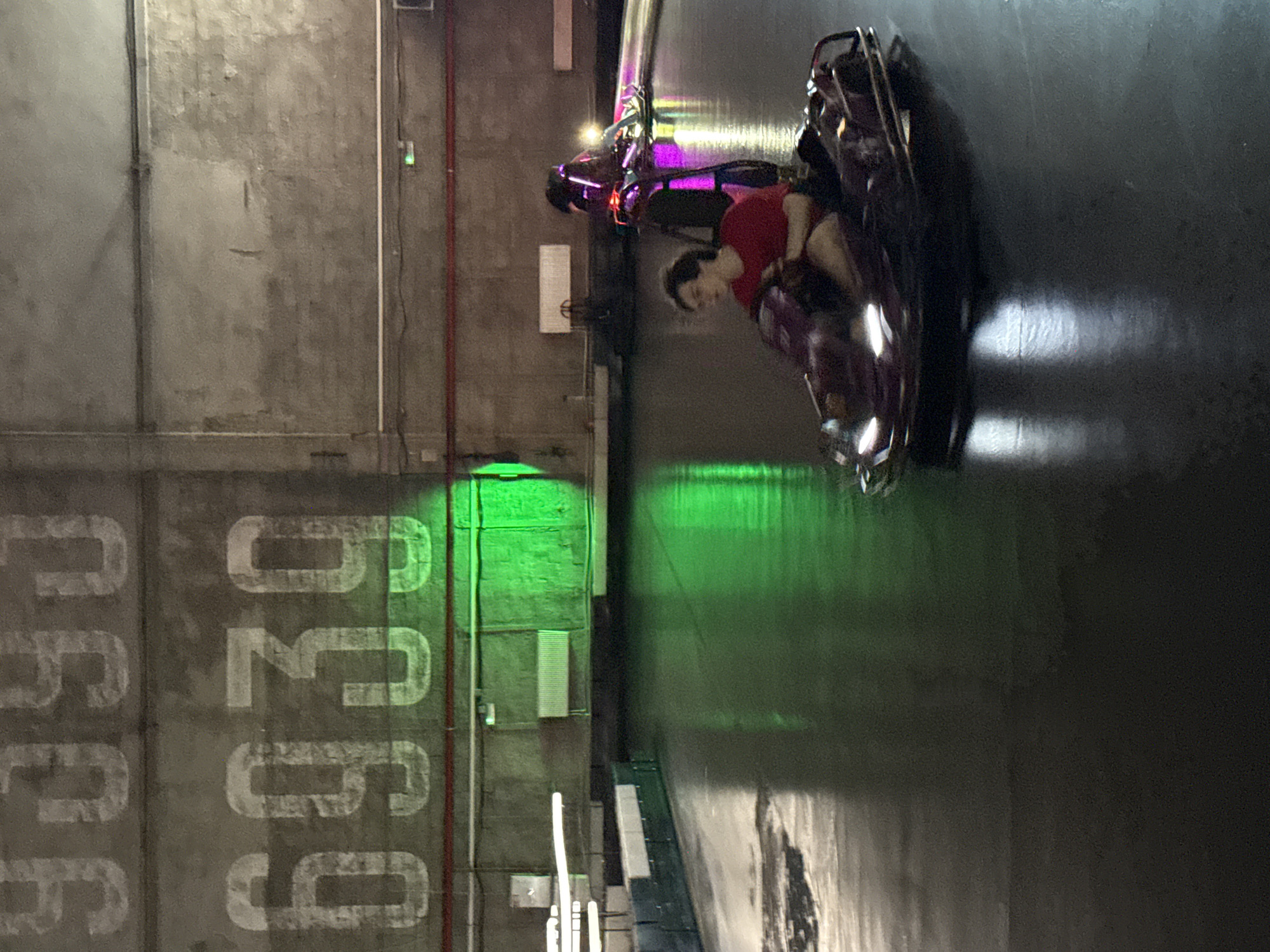 The image shows an indoor amusement park with bumper cars. Two people are visible, one driving a red bumper car and another in a purple bumper car. The environment is dimly lit with green and pink lights reflecting on the floor, suggesting a fun, energetic atmosphere. The walls have large numbers painted on them, possibly for decoration or identification. The overall impression is that of a recreational setting, likely a go-kart or bumper car arena. The specific location in Saint Petersburg, Russia is not discernible from the visual information provided in the image.