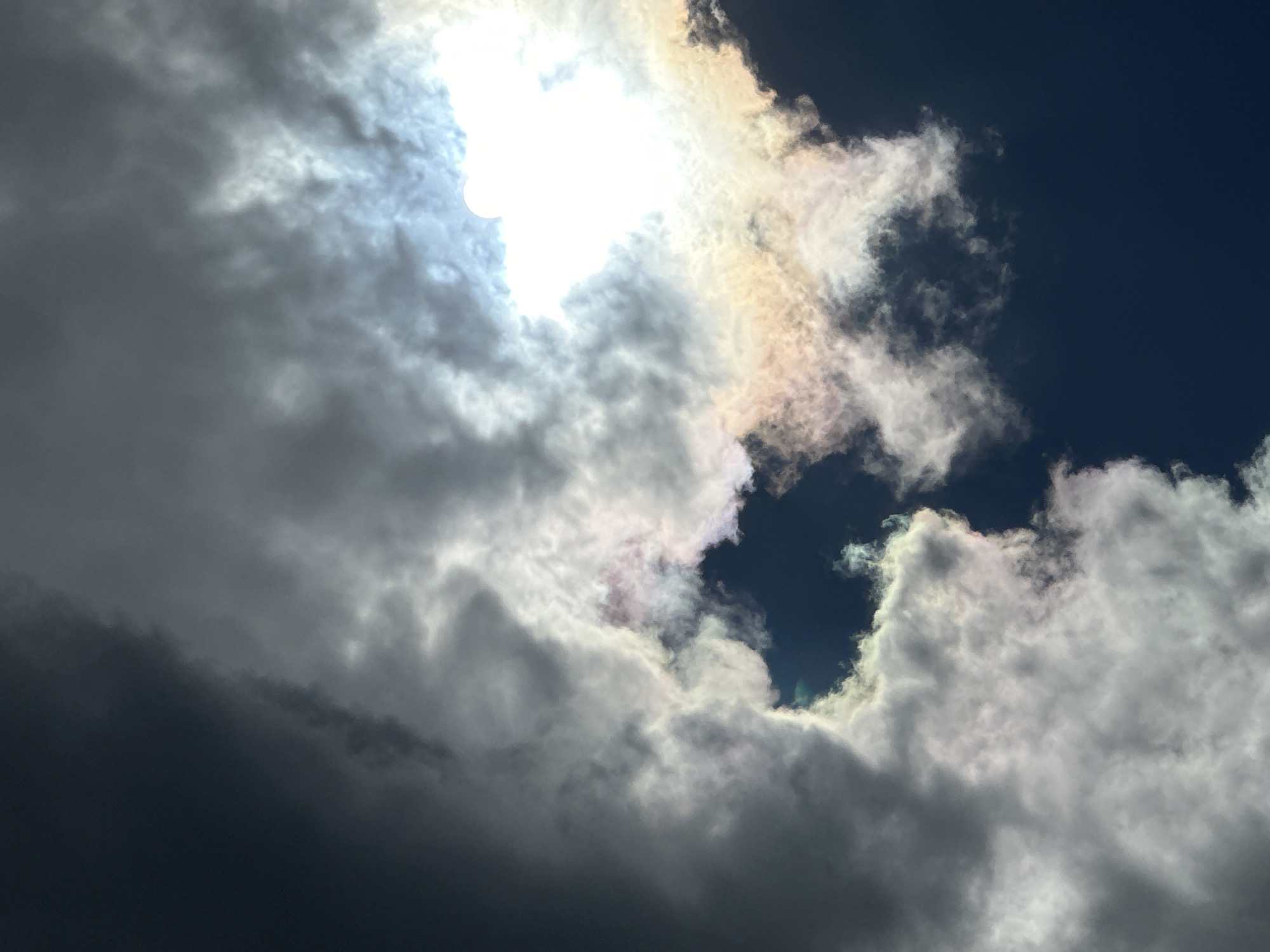 This is a low-angle, full-frame photograph of the sky. The sky is filled with dark and light gray clouds with a patch of bright sunlight peeking through. There is a faint iridescence in the clouds near the sunlight. The lower portion of the image is dark blue, indicating the sky. No people or man-made objects are visible. The location is Unknown, Unknown.