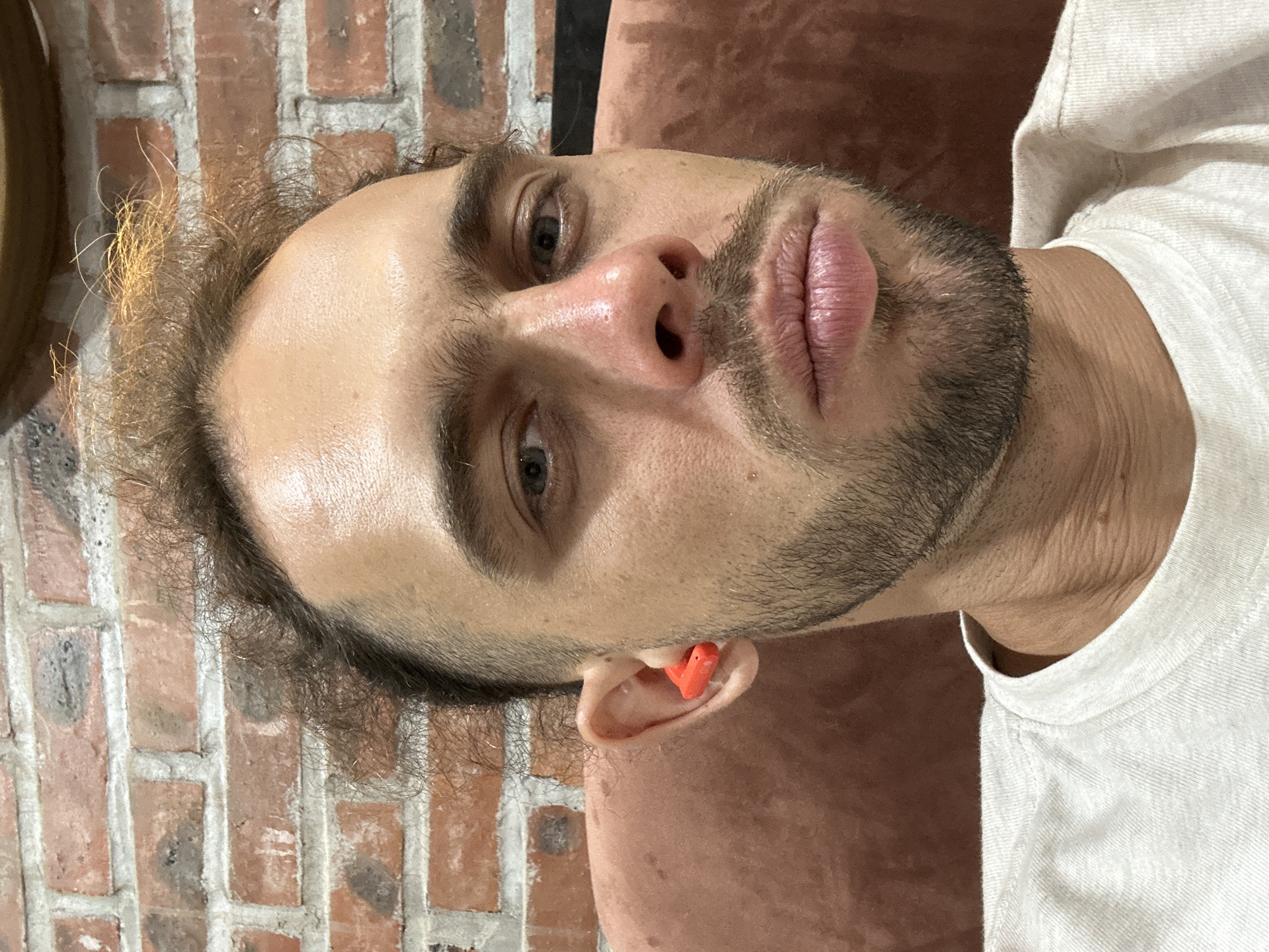 A man with a light beard and curly brown hair is looking directly at the camera. He is wearing a white t-shirt. An orange earplug is visible in his left ear. The background consists of a brick wall and a dark reddish-brown surface, possibly furniture. The image is a close-up shot of the man's face and upper body.