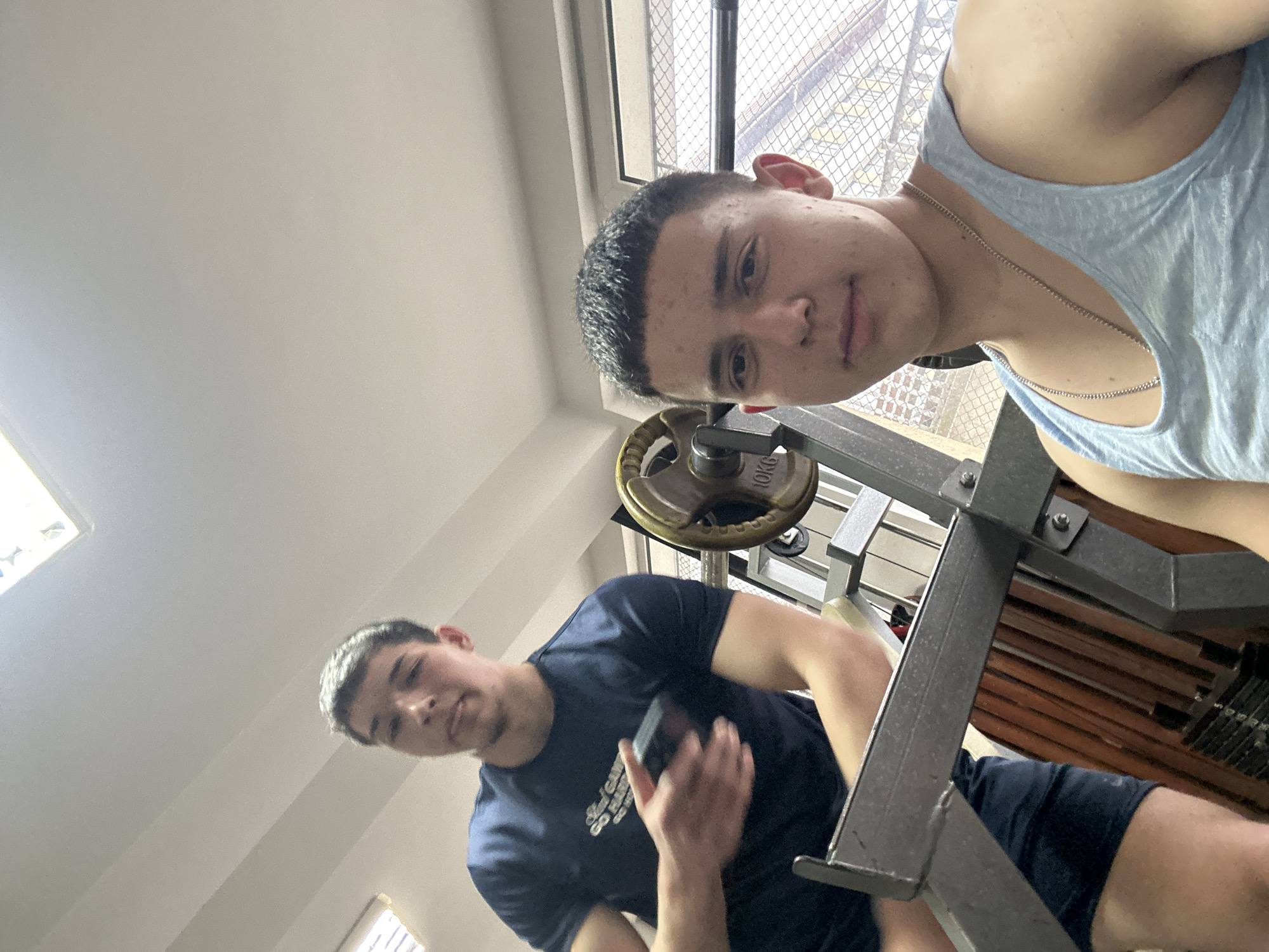 The image displays two young men indoors, likely in a gym or home gym setup in Zlatograd, Bulgaria, captured from a low, upward-looking angle.
The man in the foreground is positioned close to the camera, wearing a light blue tank top and a silver chain. He looks directly at the camera with a neutral expression, his face showing a slight sheen. Behind him, a window with a metal grate or mesh is visible.
Further back and slightly below, the second man wears a dark blue t-shirt with "Steel City" partially visible on it, and dark shorts. He looks towards the camera with a slight smirk while holding a small, black rectangular object, possibly a phone, in his right hand.
Between the two individuals and surrounding them is weightlifting equipment, including a metal frame and a visible 10KG weight plate. The ceiling is light-colored, with a light source or skylight on the left. The scene suggests the men are either taking a break from or posing during a workout session during the daytime.