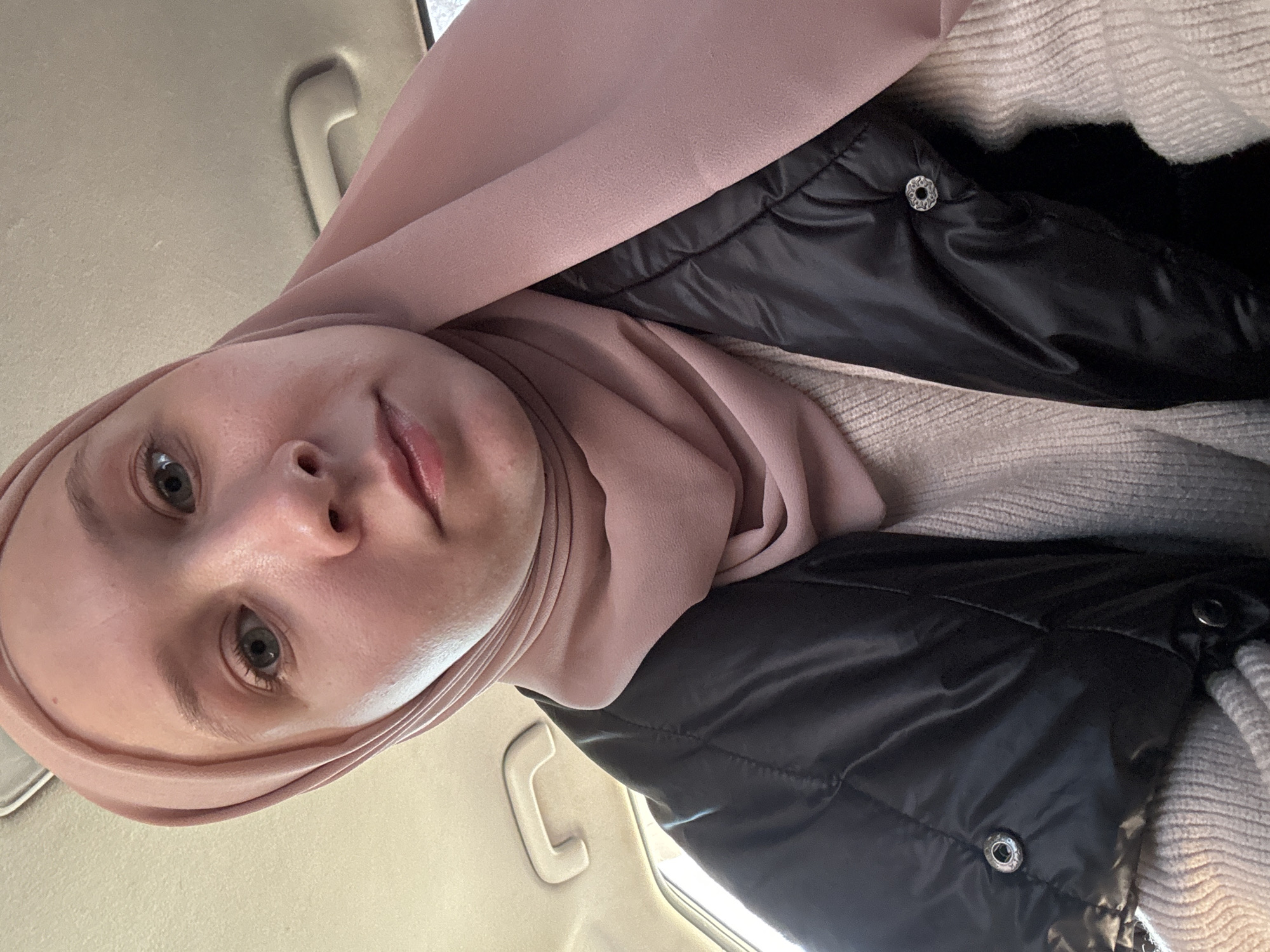 A person wearing a pink hijab and a black puffer vest over a light-colored knit sweater is inside a vehicle. The interior of the vehicle is visible, including the ceiling and a handle. The location is unknown, and there is no evidence to suggest it is Chaikovsky, Russia.