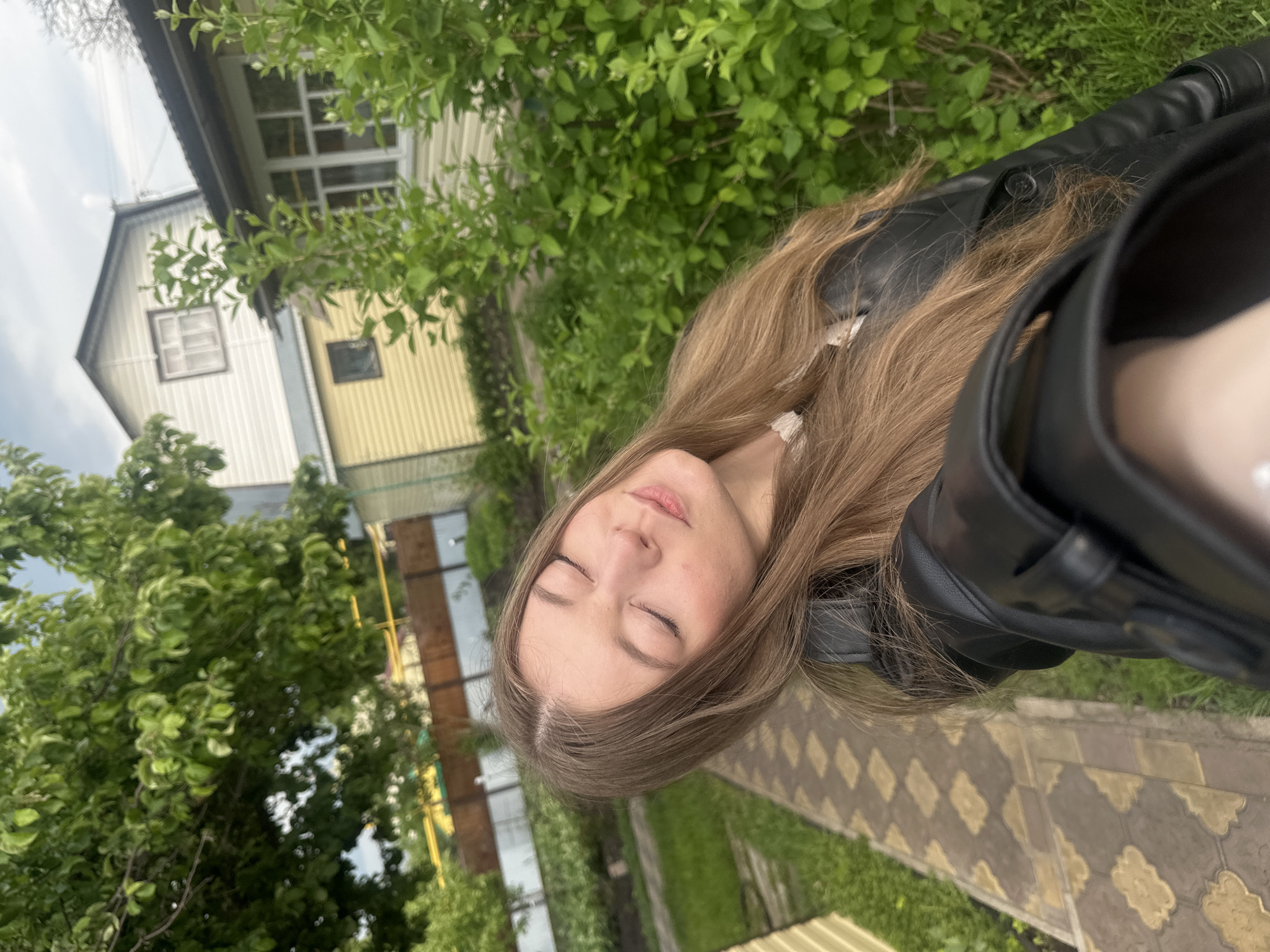 A medium-shot, slightly angled outdoor photograph shows a woman with long, light brown hair and fair skin, wearing a black leather jacket, with her eyes closed.
The background includes a house with a light yellow and white exterior, green trees and foliage, and a paved walkway with a diamond pattern. The location is plausibly Коноваловка, Russia, based on the architectural style of the house.