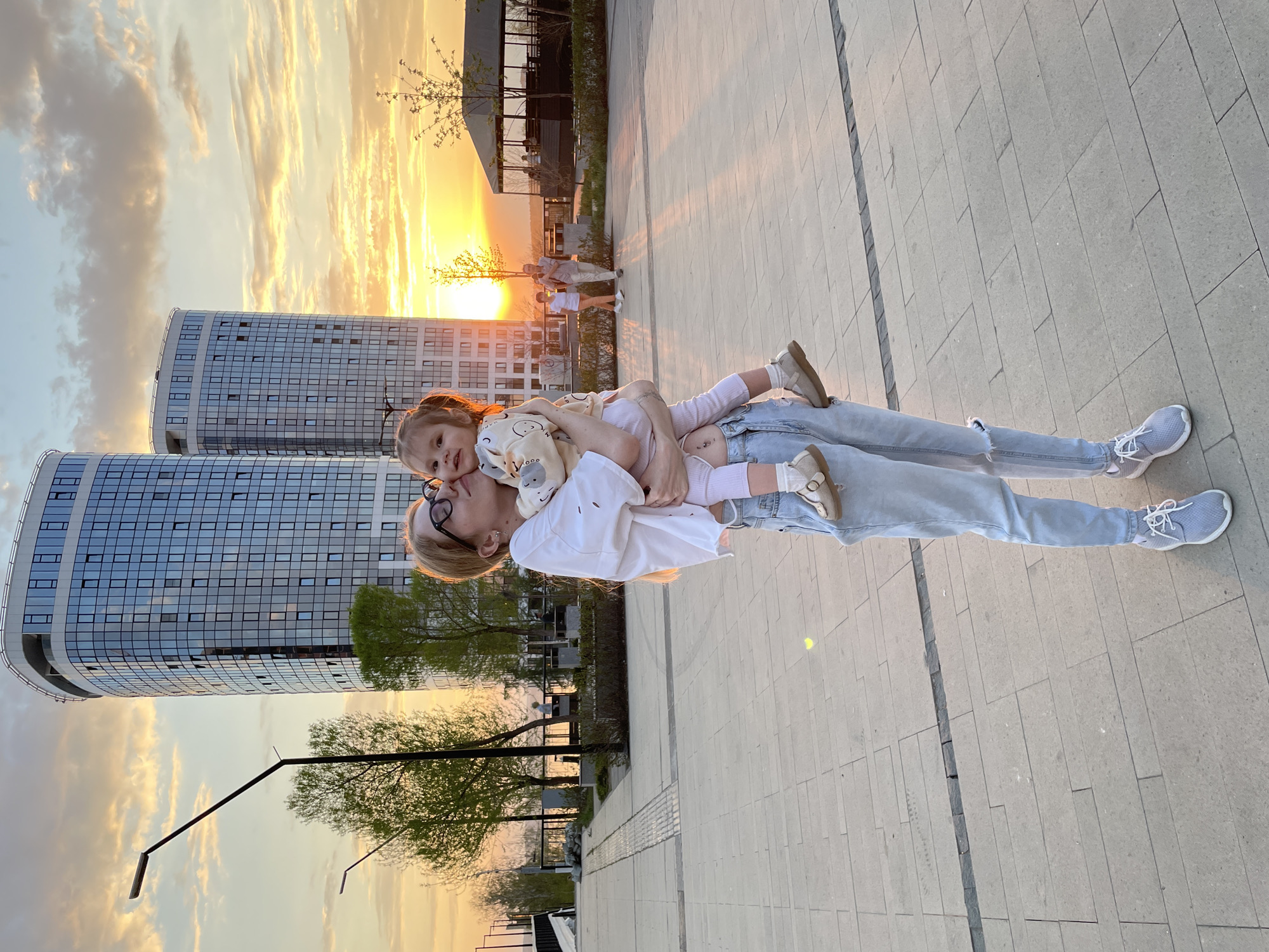 A woman with short blonde hair and glasses lies on her back on a paved plaza, holding a young girl in her arms. The girl, with light brown hair, is wearing a light-colored patterned shirt and white tights with brown shoes. The woman is wearing a white t-shirt, light-blue jeans, and gray sneakers. They appear to be interacting affectionately, possibly hugging or kissing.
The plaza is paved with light-gray rectangular stones. In the background, two modern high-rise buildings with glass facades are visible. The sky is filled with clouds and the setting sun creates a warm golden light. Trees and streetlights line the edge of the plaza. In the distance, other people can be seen walking. The setting indicates a public space, possibly a park or promenade within the city of Izhevsk, Russia.