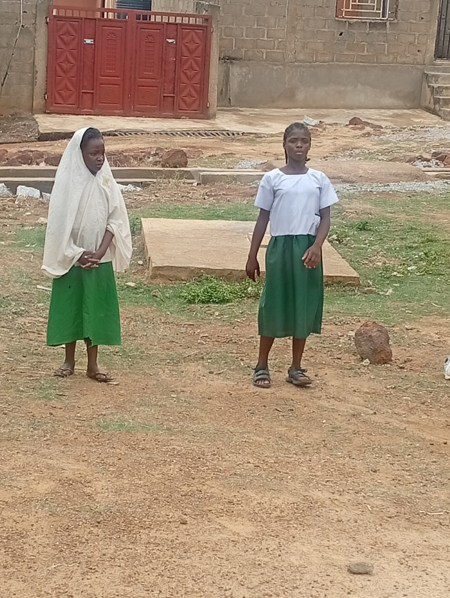 The image features two young girls standing outdoors on a dry, dirt path, consistent with a residential or semi-rural setting in Narayi, Nigeria. The time of day is daytime, with soft, even lighting suggesting either an overcast sky or bright ambient light.
On the left, a girl wears a white head covering (possibly a hijab or veil) over a white top and a long green skirt, with her hands clasped together in front of her. She is wearing sandals. To her right, another girl is dressed in a light-colored, short-sleeved top and a matching long green skirt, also wearing sandals. Both girls have neutral expressions and appear to be standing still, possibly posing for the camera. Their matching green skirts suggest they might be wearing school uniforms.
The ground around them is dusty with scattered small rocks and sparse patches of green grass. In the background, behind the girl on the left, stands a prominent reddish-brown metal gate with decorative patterns. To the right, behind the second girl, is a building constructed from unplastered concrete blocks, featuring a metal-grated window and visible steps on its far right side. Low concrete slabs or foundations are also visible on the ground behind the girls. There is no specific activity taking place, and no visible text in the image.
