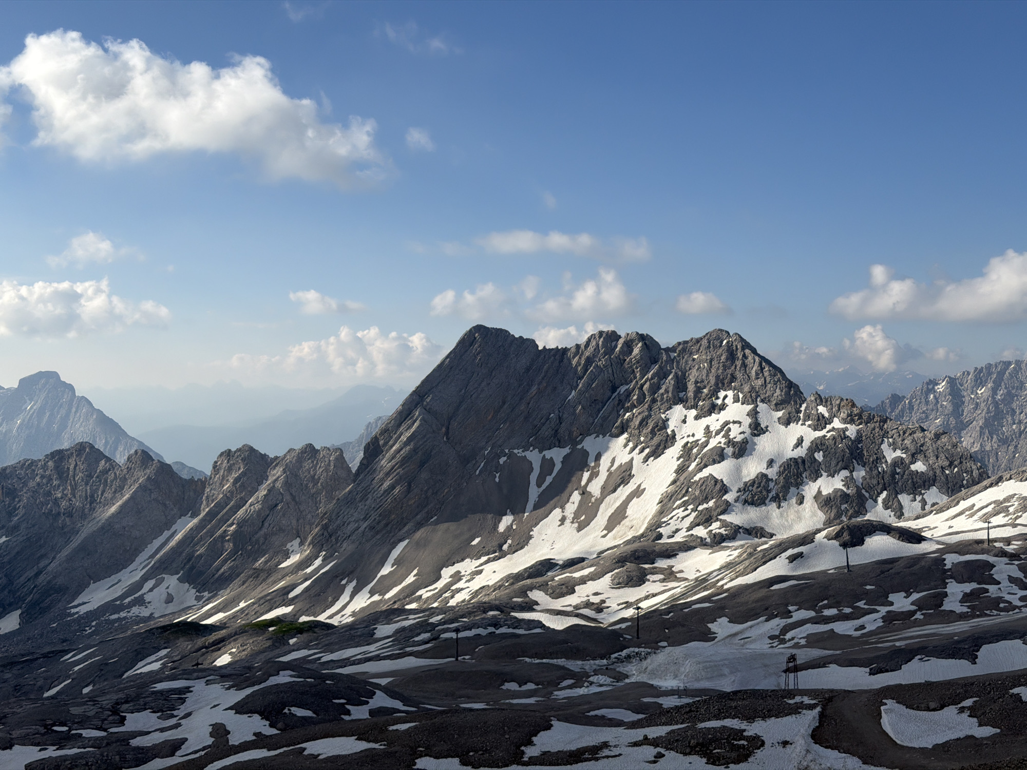 The image shows a vast mountain landscape with jagged peaks and patches of snow. The sky is a bright blue with scattered white clouds. The mountains are rocky and appear to be of a significant elevation. The foreground is covered in a mix of dark rock and melting snow, creating a textured, uneven surface. There are some man-made structures visible, including what appear to be ski lift poles and a pathway. The overall scene conveys a sense of rugged natural beauty and high altitude. This view is likely from the Zugspitze, Germany's highest peak, located near Garmisch-Partenkirchen.