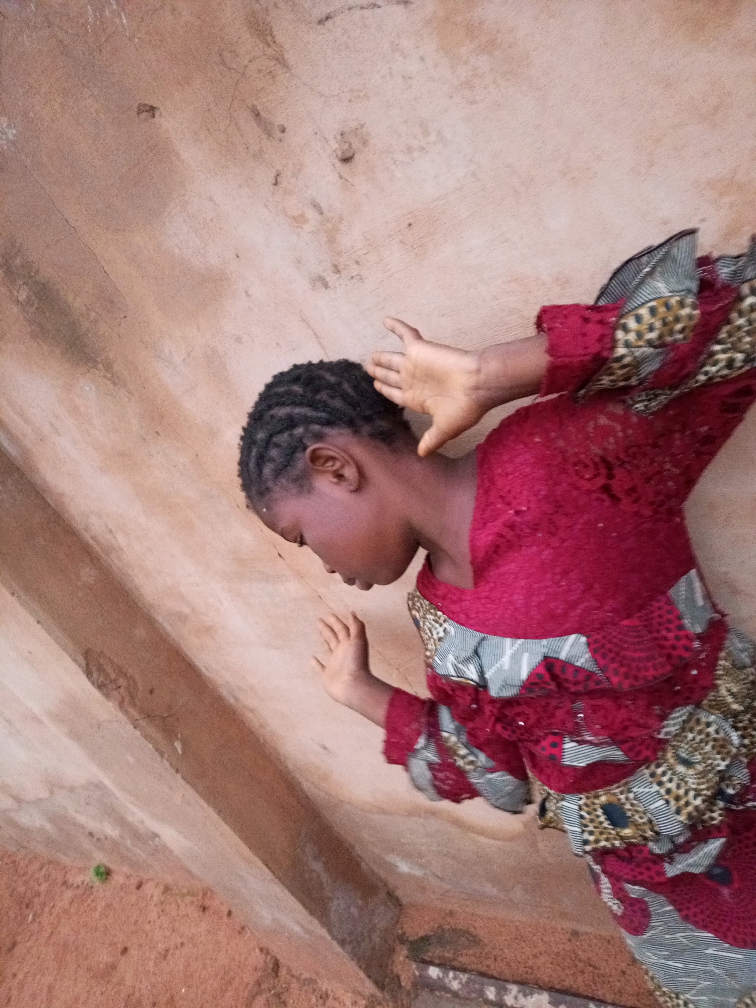 In Tumfure, Nigeria, a young individual is captured in a posed photograph. They are standing next to a textured wall, with their head tilted downwards and their eyes closed. Their hands are raised, with one resting on the back of their head and the other extended slightly forward. The individual is wearing a vibrantly patterned dress, predominantly in shades of red, black, and white, with leopard print accents. The dress features intricate detailing, including lace and ruffled layers. The background consists of a weathered, light brown wall with visible cracks and imperfections, suggesting an outdoor or semi-outdoor setting. The ground below is a reddish-brown earthy material, with hints of green foliage visible on the left side of the frame. The lighting suggests it is daytime, with soft shadows indicating a diffused light source. The overall impression is of a portrait with a slightly melancholic or contemplative mood.