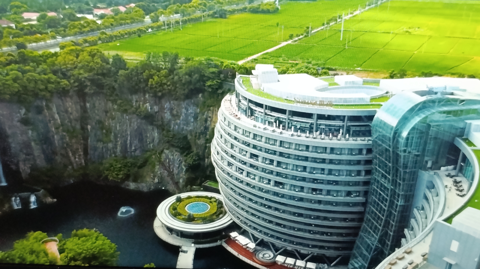 The image depicts a large, modern, multi-story building complex situated next to a sheer rock face and a body of water. The building has a distinctive curved, cylindrical shape with numerous horizontal windows and balconies. A prominent glass-fronted section juts out from the main structure. Green roofs and landscaping are visible on multiple levels of the building. In front of the building, at water level, are circular platforms with landscaping and a small pool. The body of water appears dark and reflects the surroundings. To the left of the building, a waterfall cascades down the rock face. Beyond the building and rock face, there is a vast expanse of vibrant green agricultural fields, with some roads and scattered trees. In the distance, a line of trees and buildings suggests a more developed area or a town. The sky is not clearly visible. The overall scene suggests a resort or hotel designed to integrate with the natural environment. This building is the InterContinental Shanghai Wonderland Hotel, located in China, not Moldova.