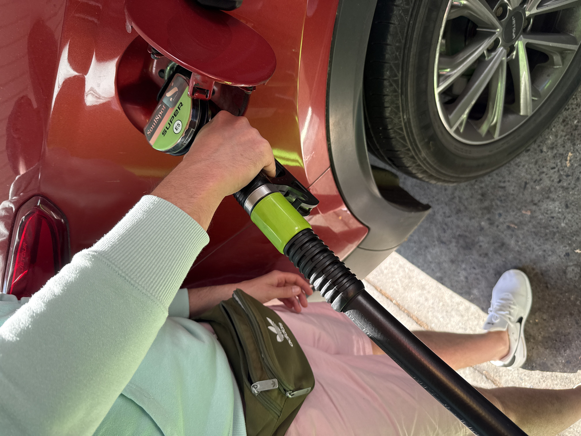 A person is filling a red car with fuel from a green gas pump nozzle at a gas station, likely in Bad Lauterberg im Harz, Germany. The person is wearing a light blue sweatshirt, pink shorts, white sneakers, and a green fanny pack with an Adidas logo. The gas pump nozzle is black with a bright green handle. The car's fuel cap is open, revealing a "SoulSpot SUPER E5" label. The car's tire and wheel are visible in the foreground, and the ground appears to be paved. The lighting suggests it is daytime.