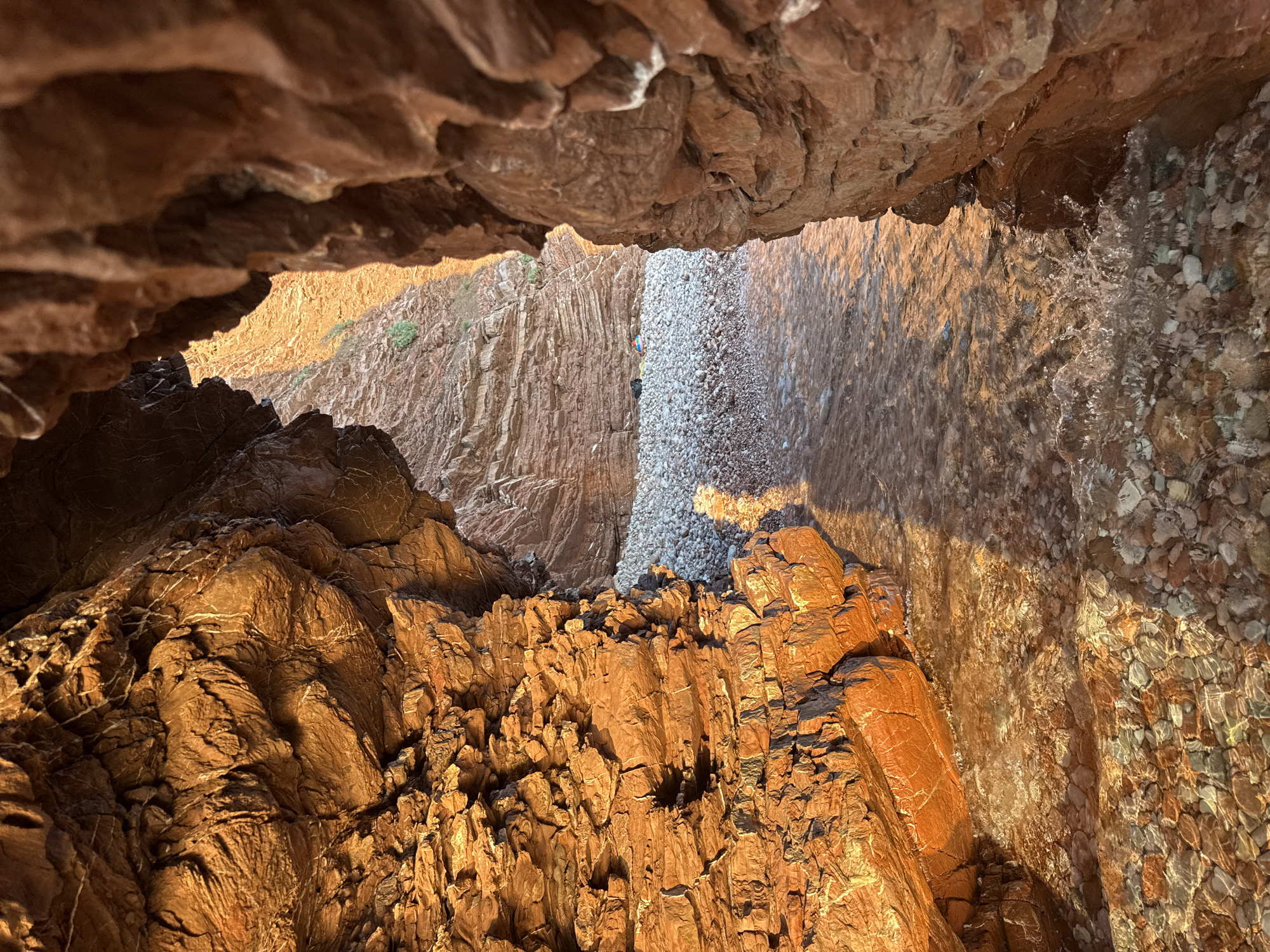 The image captures a narrow passage between towering, striated, reddish-brown rock formations, likely within a sea cave or a naturally formed canyon near Sveti Stefan, Montenegro. The rock faces are rough and textured, catching the warm, golden light of what appears to be either sunrise or sunset. The light illuminates the layers within the rock and casts a warm glow on the scene.
At the end of the passage, a small, pebbly beach is visible, with smooth, rounded stones in various shades of grey, white, and reddish-brown. The water is clear and shallow, with gentle ripples and reflections of the warm light on its surface. The sea floor beneath the water is also covered in pebbles. In the distance, on the beach, a small bundle of items, possibly a bag and a towel, are visible, suggesting a solitary visitor or prior presence. The overall impression is one of a secluded and serene natural environment.