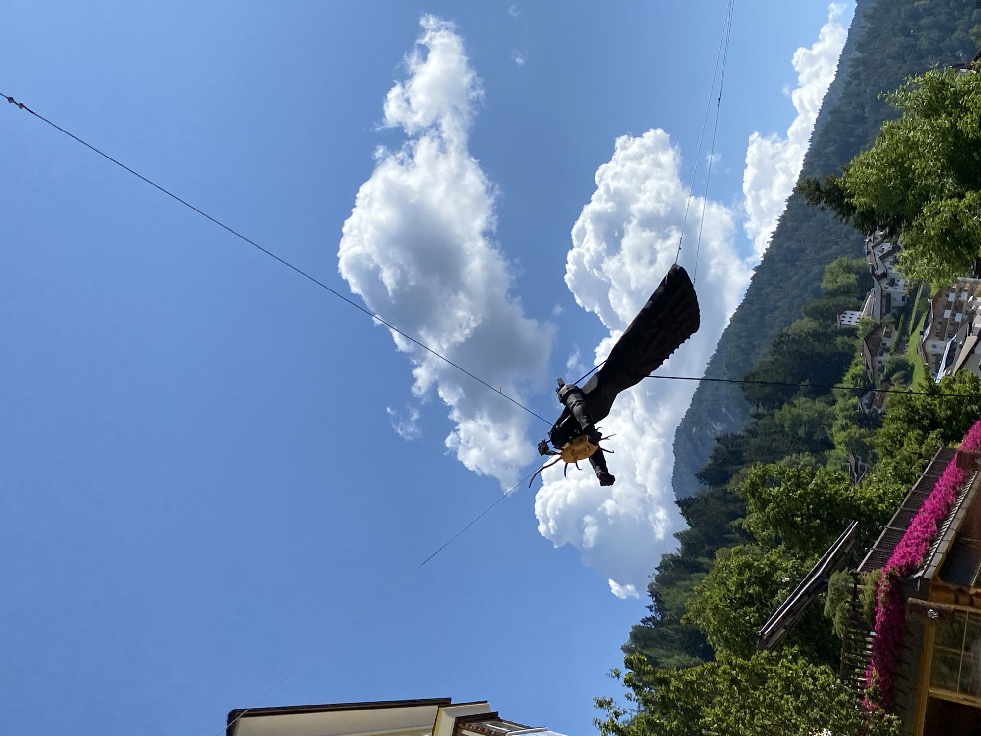In Urtijëi - St. Ulrich in Gröden - Ortisei, Italy, a sun-like effigy with spiky rays, possibly part of a local festival or decoration, is suspended from wires against a bright blue sky dotted with fluffy white clouds. The effigy appears to be made of dark material, possibly fabric or metal, with a yellow or golden face. Below the effigy, a steep, tree-covered mountainside is visible, dotted with traditional alpine houses. In the foreground, the upper portion of a building with a balcony adorned with vibrant pink flowers can be seen. The overall impression is a festive, outdoor scene in a mountainous, European town.