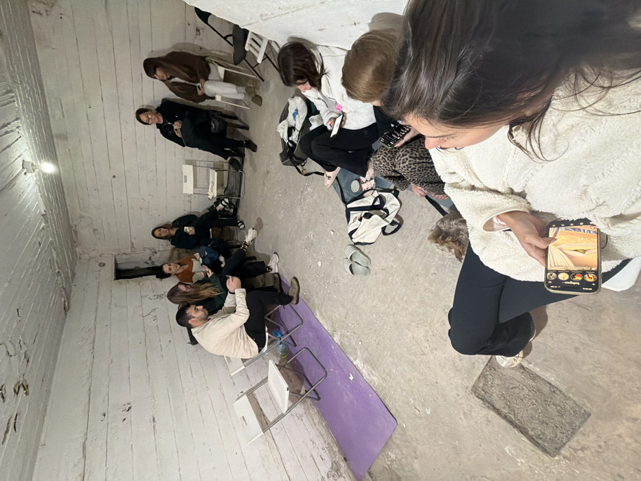 The image shows approximately ten individuals gathered in a concrete bomb shelter located in Tel-Aviv, Israel. The room features rough, unpainted concrete walls with horizontal striations, painted off-white, and a similarly textured concrete ceiling with a single exposed light bulb for illumination. The floor is bare concrete. A dark, rectangular opening is visible in the upper section of the back wall.
Seven individuals are seated on white plastic folding chairs, arranged in a semi-circle. Many are focused on their mobile phones; for example, a man in a beige sweatshirt and dark pants on the left is looking at a phone, as is a woman beside him in a green top.
In the foreground, three additional individuals are positioned on the concrete floor. One person, viewed from the shoulder and back, holds a smartphone displaying a social media application with an image of an interior room. A small, light-brown furry dog is partially visible next to this individual. A purple yoga mat is laid on the floor. Nearby, a pair of light-colored slippers and a dark duffel bag containing clothing items are present. The scene depicts a group in a state of casual waiting or temporary refuge.
