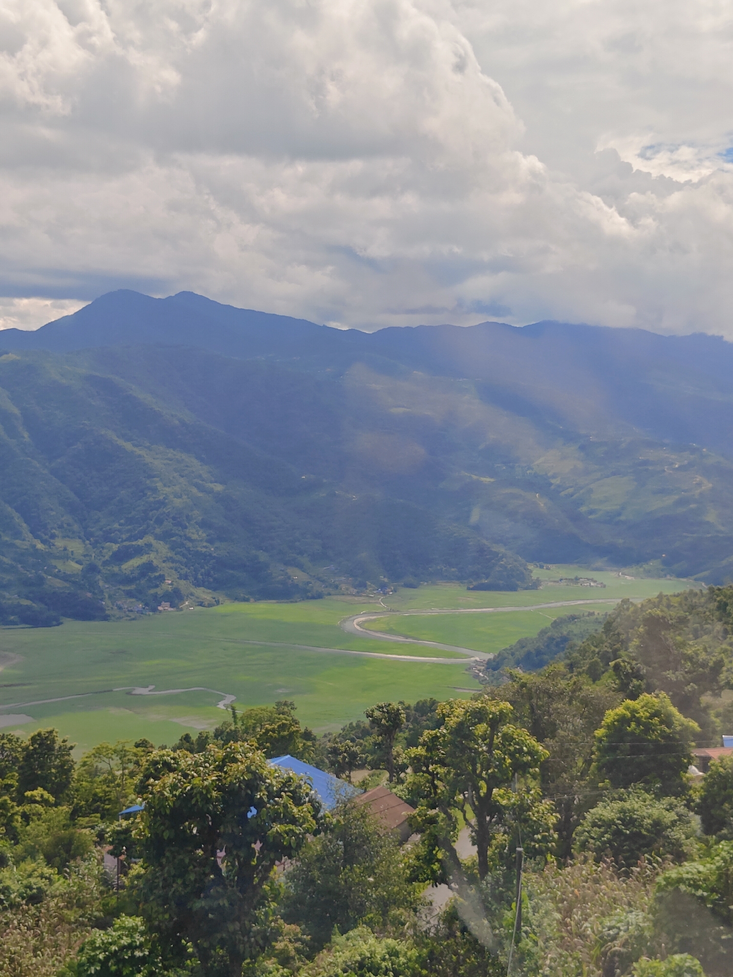 This is an image of a vast green valley with a winding river, framed by lush, tree-covered hills and majestic mountains in the distance. The sky is filled with dramatic clouds, with sunlight breaking through in some areas, creating shafts of light over the landscape. In the foreground, dense vegetation and trees are visible, with a hint of rooftops, suggesting a settlement nestled within the hills. The scene appears to be taken from an elevated viewpoint, offering a panoramic vista of the natural beauty of the area. This landscape is characteristic of the outskirts of Pokhara, Nepal, likely a region known for its verdant valleys, rivers, and proximity to the Annapurna mountain range, contributing to its scenic appeal. There are no people or specific actions depicted in the image.