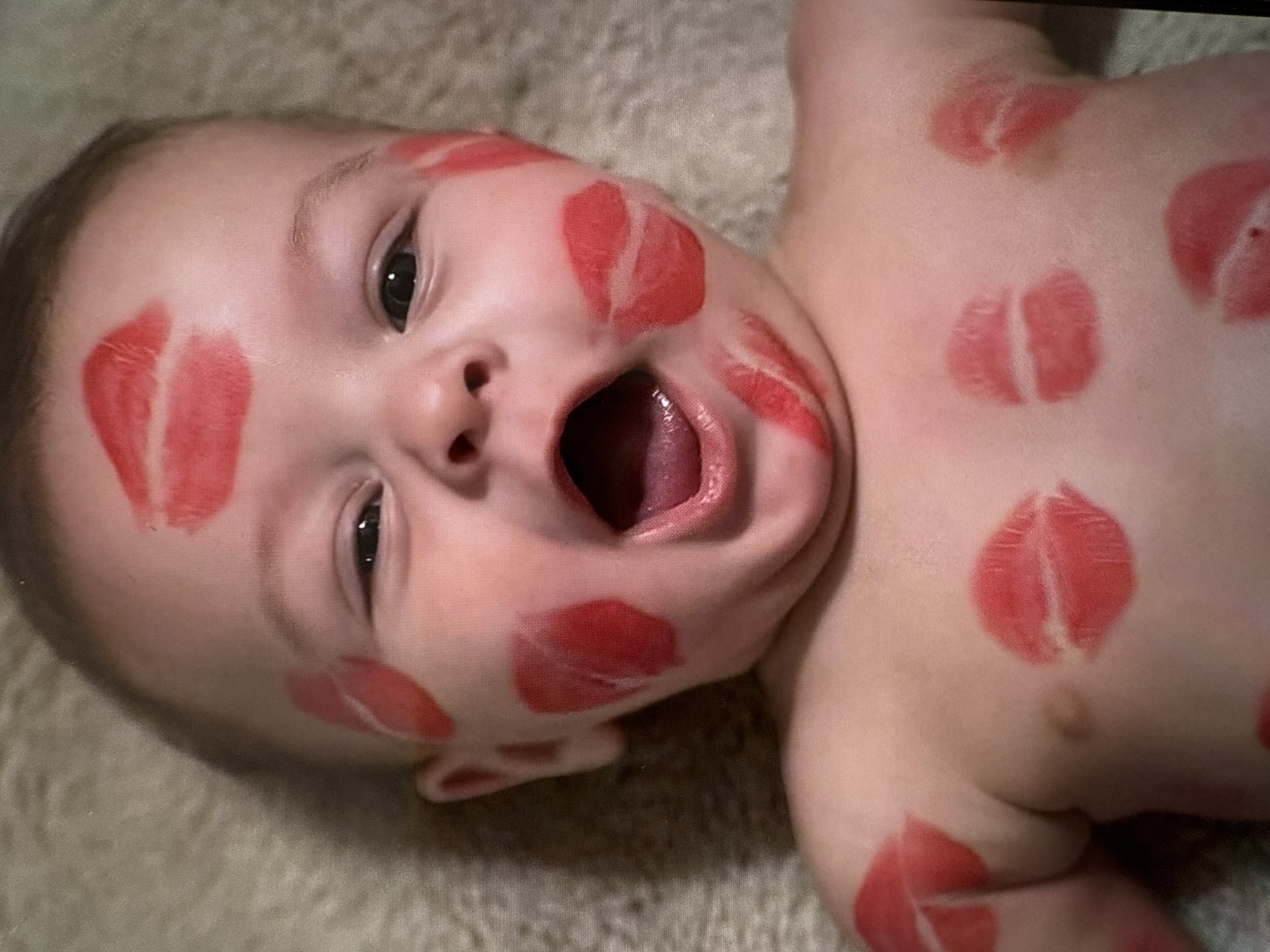 The image presents a close-up, overhead view of an infant with fair skin, dark eyes, and light brown hair, depicted in a supine position. The infant's mouth is wide open, revealing the tongue, suggesting an expression of laughter or vocalization, with eyes wide and directed towards the viewer. The face, shoulders, and upper chest are adorned with numerous vibrant red lipstick impressions, simulating kiss marks. These marks are visible on the forehead, both cheeks, chin, left shoulder, and multiple locations across the chest, varying in shape and intensity. The infant appears to be bare-chested. The background consists of a uniformly textured, light beige or off-white soft material, consistent with a blanket or rug. The image itself contains no discernible visual elements or contextual information that would allow for the identification of its specific location, such as the city of Budva, Montenegro.
