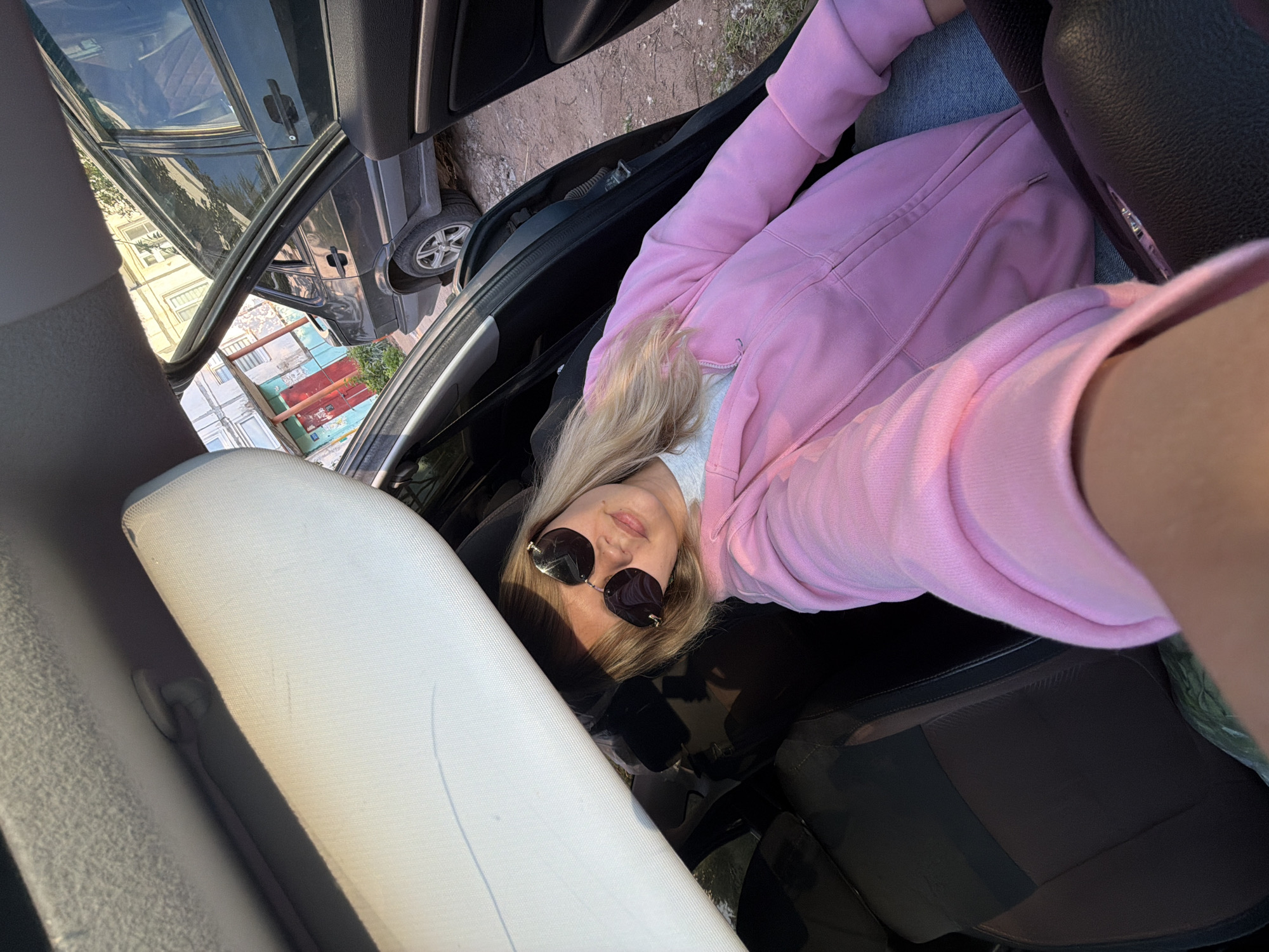 A bright daytime selfie captures a blonde woman seated inside a car with an open door, looking directly at the camera. She wears a light pink hooded sweatshirt over a lighter top and blue jeans, with large, dark sunglasses reflecting the bright environment. Her blonde hair frames her face and shoulders.
The perspective is from within the car, looking out through the open driver's or passenger's door. The car's interior, including parts of a dark dashboard, a dark seat, and a light-colored headrest, is visible. Outside the car, another dark grey or black vehicle is partially parked, with its front wheel visible.
In the background, beyond the vehicles, a street or ground area transitions to multi-story residential buildings. One building stands out with distinct painted sections in red, blue, and white. Trees and foliage are scattered between the buildings.
The bright sunlight and strong reflections indicate a clear, sunny day, likely late morning or afternoon. The setting is an urban or suburban residential area in Chaikovsky, Russia. No visible text is present.