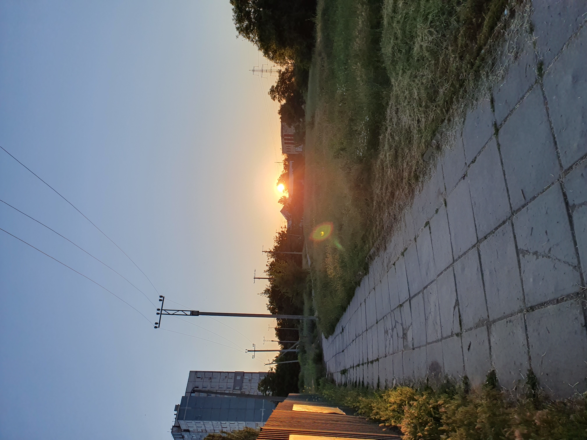A concrete sidewalk runs from the bottom left towards the middle right of the frame, flanked by a tall, thin, corrugated metal fence on the left and a grassy, overgrown field on the right. In the distance, several utility poles with wires stretch across the sky, which is a clear, pale blue transitioning to a warm orange glow near the horizon. A tall, grey apartment building stands on the far left, partially obscured by trees. The setting sun is visible in the center background, peeking through trees and illuminating the sky with a warm light. The scene appears to be an urban or suburban area at dusk.