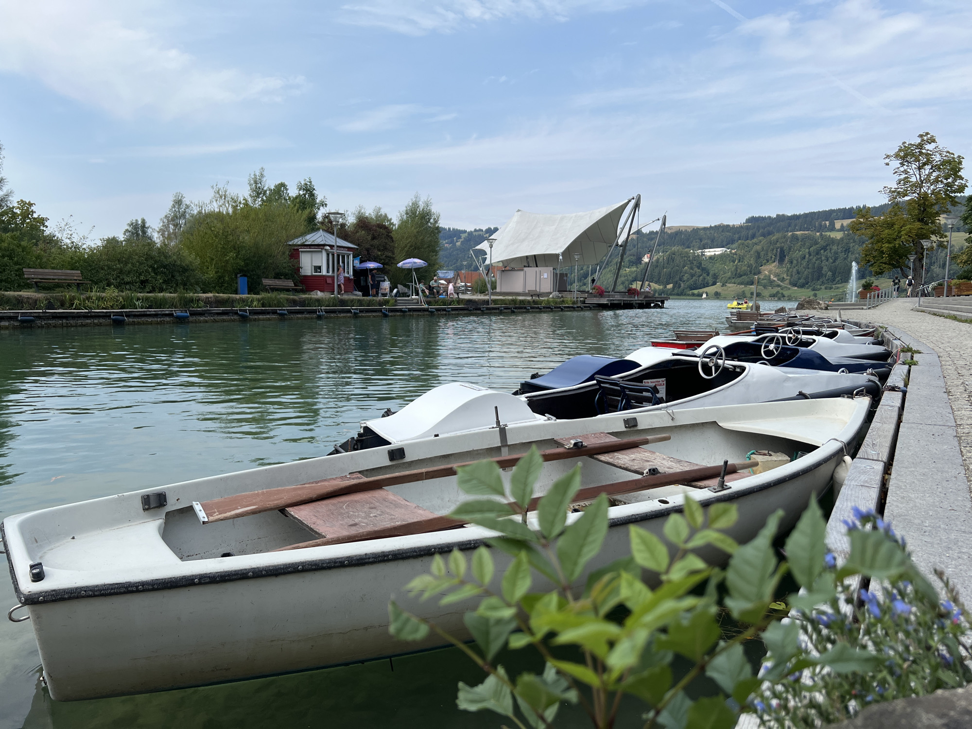 The image shows a row of small boats docked along the shore of the lake in Immenstadt im Allgäu, Germany. The boats appear to be rented paddle boats or small rowboats. In the background, there is a red building with white trim, likely a rental kiosk, and a large white tent structure. The lake is calm and the sky is partly cloudy. The shore is lined with a paved walkway and a stone wall. Lush green trees and hills are visible in the distance. There are a few people scattered around, some sitting and some walking.