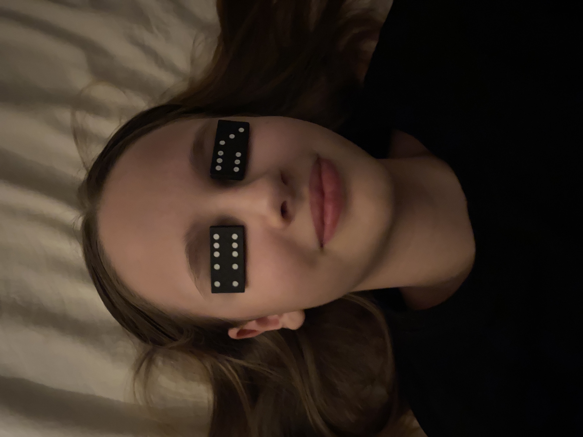 A person, likely a young female, lies supine on a wrinkled, light-colored surface, possibly a bed. Two dominoes are placed over their eyes, each displaying six dots. The person's hair is brown and spread around their head on the surface. They are wearing a dark-colored top. No location information pertaining to Yekaterinburg, Russia, is discernible from the image.