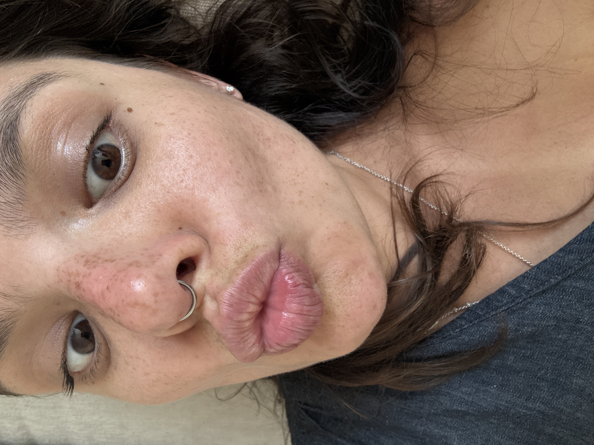 The image is a close-up portrait of an adult female subject, located in Tel-Aviv, Israel. The subject's face is prominent, featuring wide-open brown eyes; the left iris is directed towards the viewer's right, and the right iris towards the viewer's left. The lips are pursed in a 'kissing' expression, appearing full and glossy with vertical creasing. A silver-colored circular septum piercing is present. The skin displays numerous scattered freckles and several distinct moles, primarily on the right cheek. The bridge and tip of the nose exhibit reddish discoloration and small blemishes. Dark, wavy hair frames the face, and a thin silver chain necklace is visible around the neck. A small, white stud earring is present in the visible earlobe on the viewer's right. The subject wears a dark, possibly denim-textured, garment. The background is indistinct and appears to be a light, uniformly textured surface.
