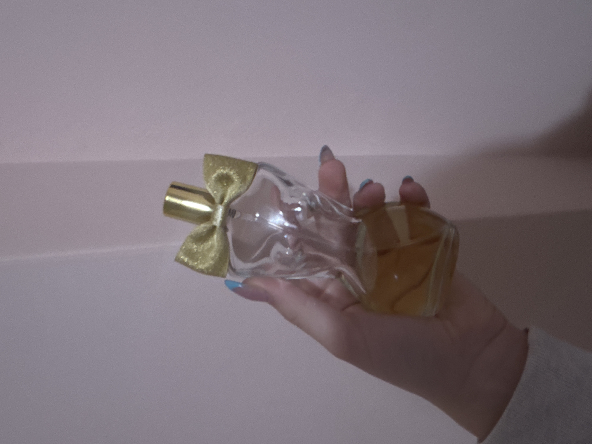 A person is holding a perfume bottle in their hand. The bottle is clear with a golden bow on the cap and a golden-colored liquid inside. The person has pale skin and their fingernails are painted with a light blue color. The person is wearing a white sweater. The background is a light purple-pink wall. The image appears to be taken indoors. There is no evidence to suggest that this image was taken in Lehchevo, Bulgaria.