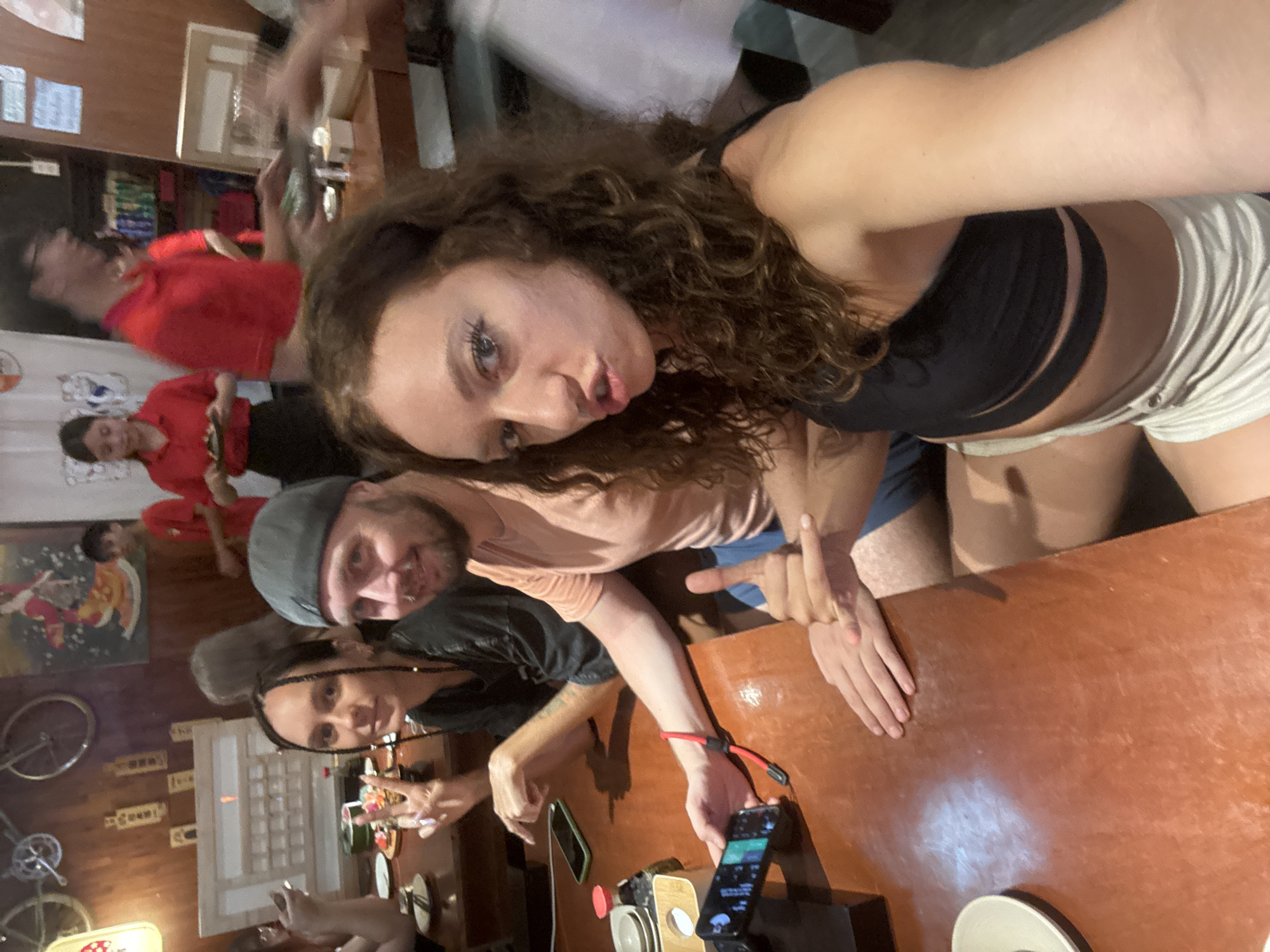 A selfie taken at a table inside a business in Đà Nẵng, Vietnam. There are 3 people visible in the front of the image and some blurry patrons in the back. The person taking the selfie is a woman with curly brown hair and a black shirt. To the left is a man with a grey beanie and a pinkish shirt. A woman with dark braids and a dark shirt is partially visible in the bottom left. There is a cell phone visible on the right side of the table and some plates, condiments, and other restaurant items. Decorations on the wall include bicycles and Japanese script. Patrons are wearing red shirts in the background of the establishment.
