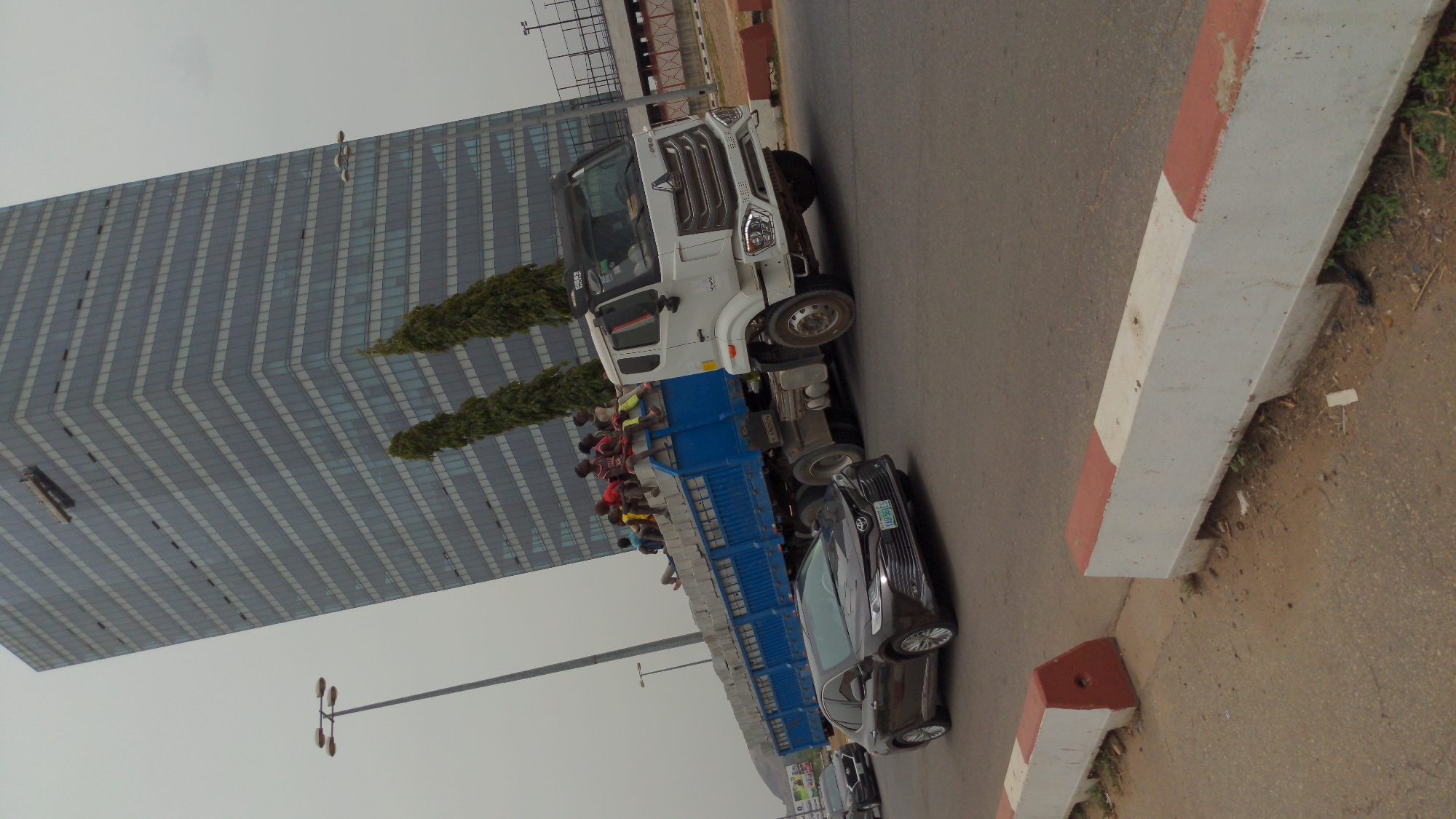 An outdoor daytime scene captures a bustling street in an urban environment, identified as Abuja, Nigeria. The image is composed from a slightly elevated vantage point, looking down along the road.
In the foreground and midground, a white and blue flatbed truck is the central focus. It is heavily loaded with numerous concrete blocks or similar building materials. Precariously seated and standing on top of these materials in the open bed of the truck are many individuals, primarily men and boys, dressed in casual attire such as t-shirts and shorts. This suggests an informal mode of transport for both people and goods.
Behind the truck, a dark-colored sedan, possibly a Toyota Camry, is visible on the road. Its license plate is partially legible, displaying "LAGOS SBB-xxxx," indicating its registration from Lagos State. Another dark vehicle is partially discernible further down the road.
To the right side of the street, a concrete curb separates the paved road from a dirt verge. This curb is distinctly painted with alternating sections of white and reddish-brown.
Dominating the left background is a large, modern skyscraper. This building features a striking facade of grey-blue glass panels arranged in a distinct diagonal or chevron pattern. In front of the skyscraper, a row of green trees or hedges adds a touch of nature to the urban landscape. A tall lamppost with multiple light fixtures stands in the midground, extending towards the sky. The sky above is overcast, indicating a cloudy day with diffused lighting and an absence of harsh shadows. The overall scene depicts everyday urban activity.