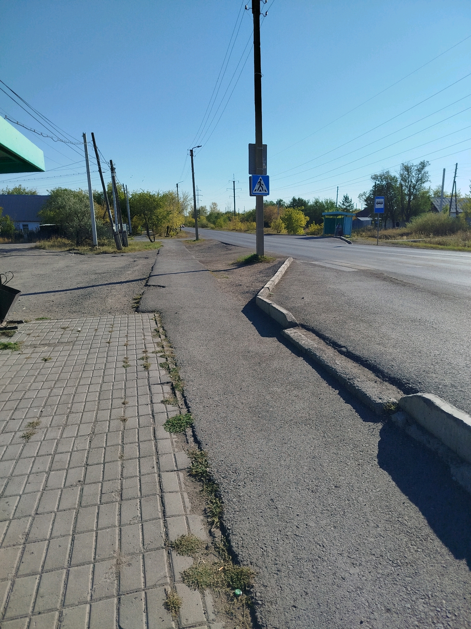 The image shows a sunny day in a suburban area with a clear blue sky. In the foreground, a paved sidewalk with grey square tiles runs along the left side of the image, bordered by asphalt. To the right of the sidewalk is a road with a concrete curb that appears to be in disrepair in places. Electrical poles with numerous wires crisscross the sky, and trees with yellowing leaves line the sides of the road. In the background, there are a few houses with dark roofs and a bus stop with a blue sign indicating a bus route. A pedestrian crossing sign is visible on a pole. The scene suggests a rural or semi-urban setting.
