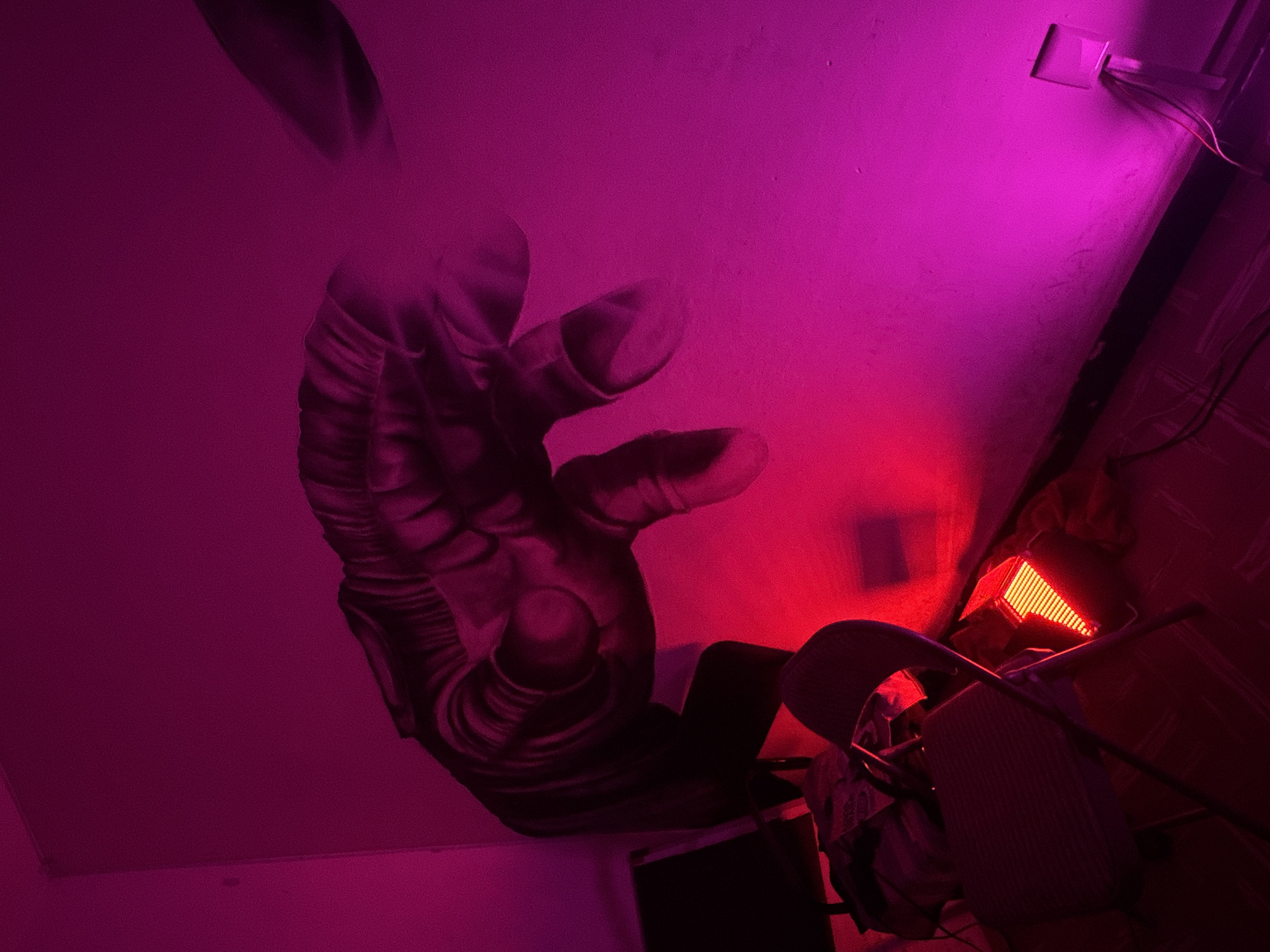 The image captures an indoor scene dominated by artificial lighting in shades of pink, magenta, and red, suggesting a nighttime or low-light setting. A large, detailed mural of a human hand is painted in grayscale tones on a light-colored wall or ceiling, appearing as a right hand with the palm facing forward and fingers slightly curled, showcasing anatomical details like veins and skin texture.
The lighting creates a distinct gradient, with magenta and pink hues illuminating the upper and left parts of the wall and mural, while an intense red light source brightens the lower right section. In this red-lit area, a metallic-framed chair with a fabric seat and back is visible, positioned at an angle. Adjacent to the chair is a rectangular light fixture, likely an LED panel, emitting a powerful red-orange glow. In the upper right corner of the image, a white electrical outlet or light switch is mounted on the wall, with wires extending from it.
No people are visible, and no activity is explicitly occurring. The scene conveys a moody or artistic atmosphere, likely a decorated room or creative space. No discernible text is present. The location context indicates this scene is in Puebla, Mexico.