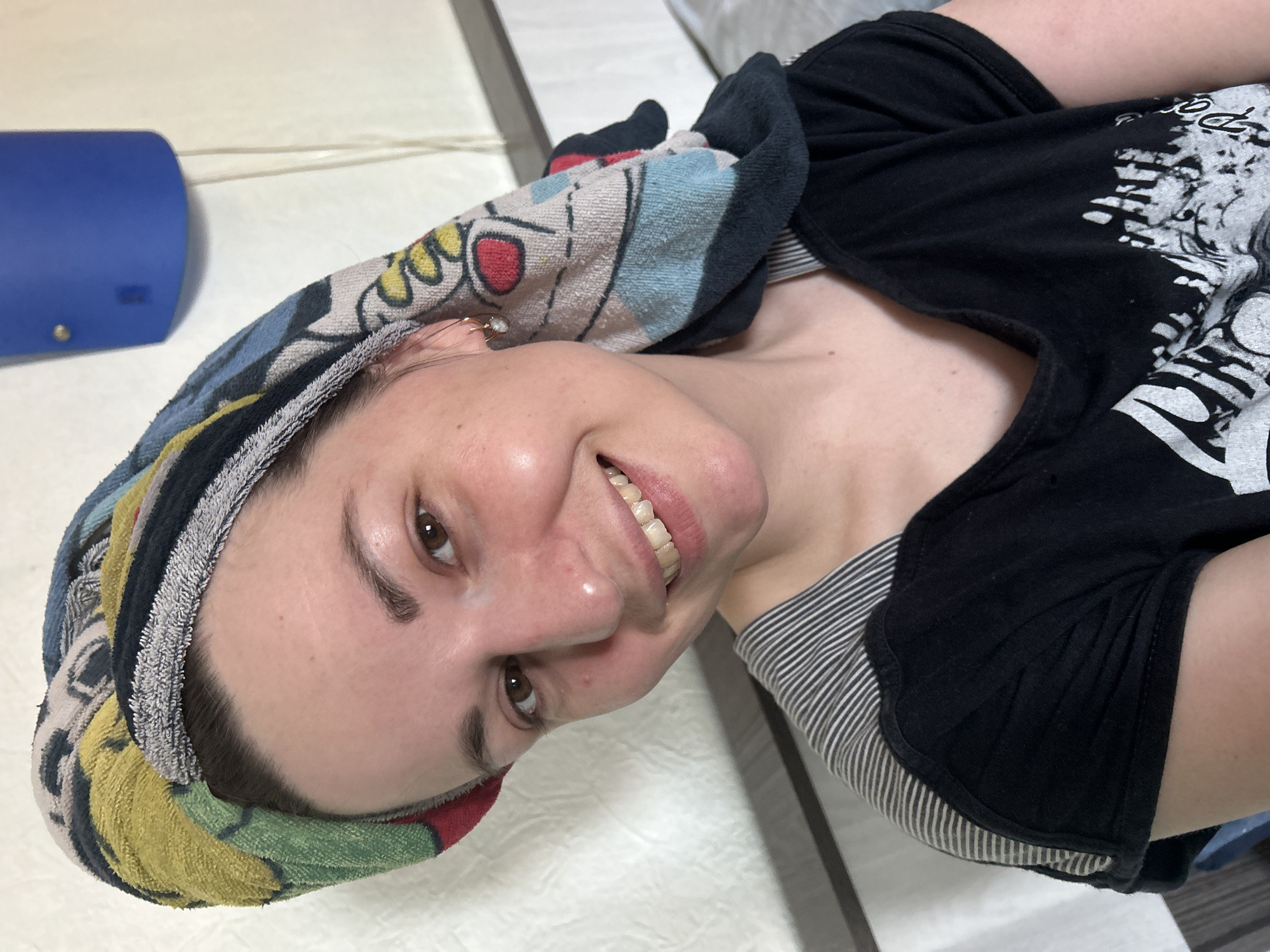 A woman is pictured indoors, smiling at the camera. She has a colorful towel wrapped around her head. She is wearing a black t-shirt over a striped top. A blue cylindrical object is visible in the background. The location is unknown.