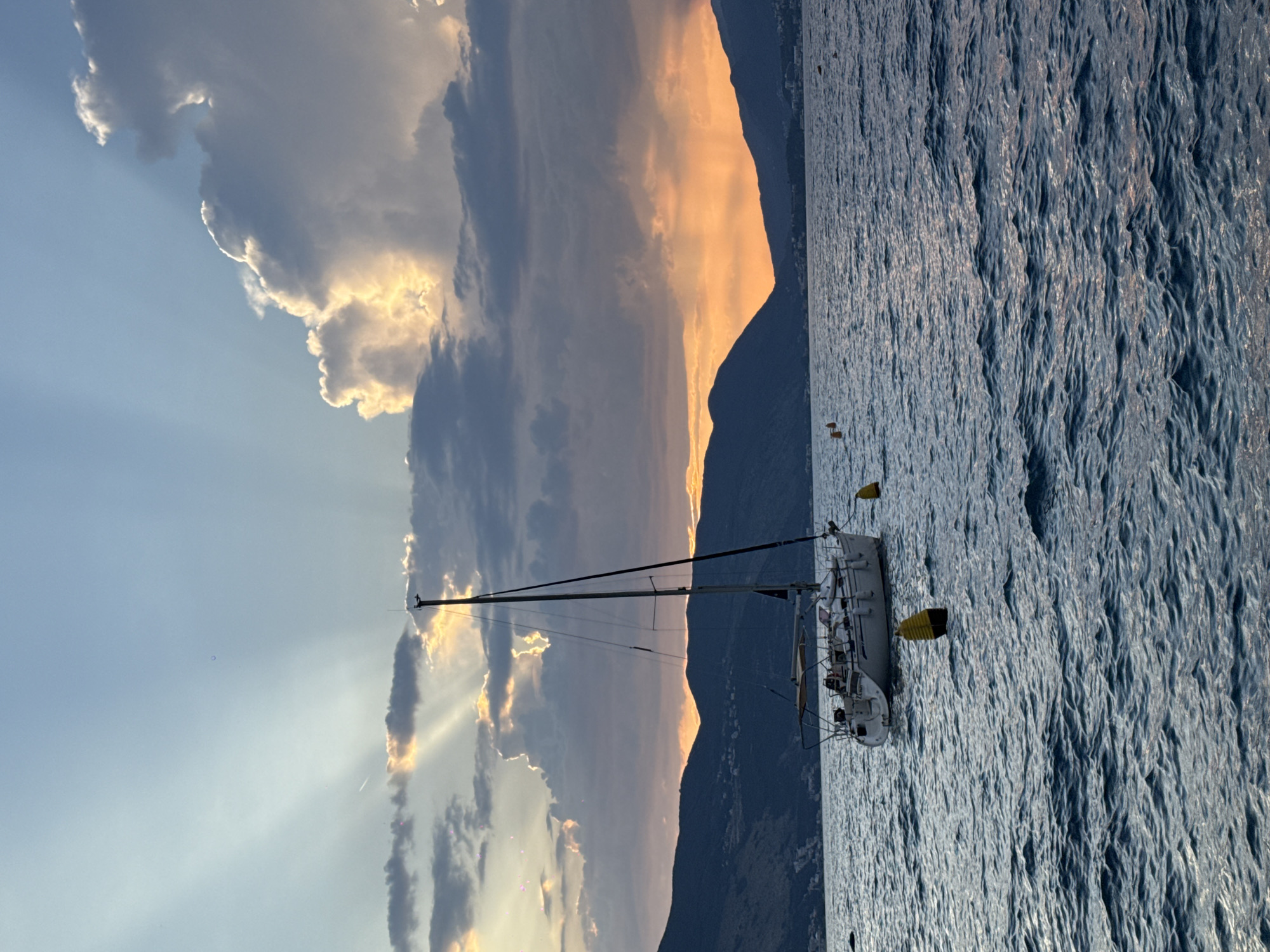 A sailboat is anchored in the water near a mountainous coastline. The sun is setting, casting an orange glow on the clouds and water. The sky is clear, with some scattered clouds. The water is calm, with gentle waves. The sailboat is white with a single mast. There are no people visible on the sailboat. The mountains are dark and silhouetted against the sunset. The overall mood of the image is peaceful and serene.