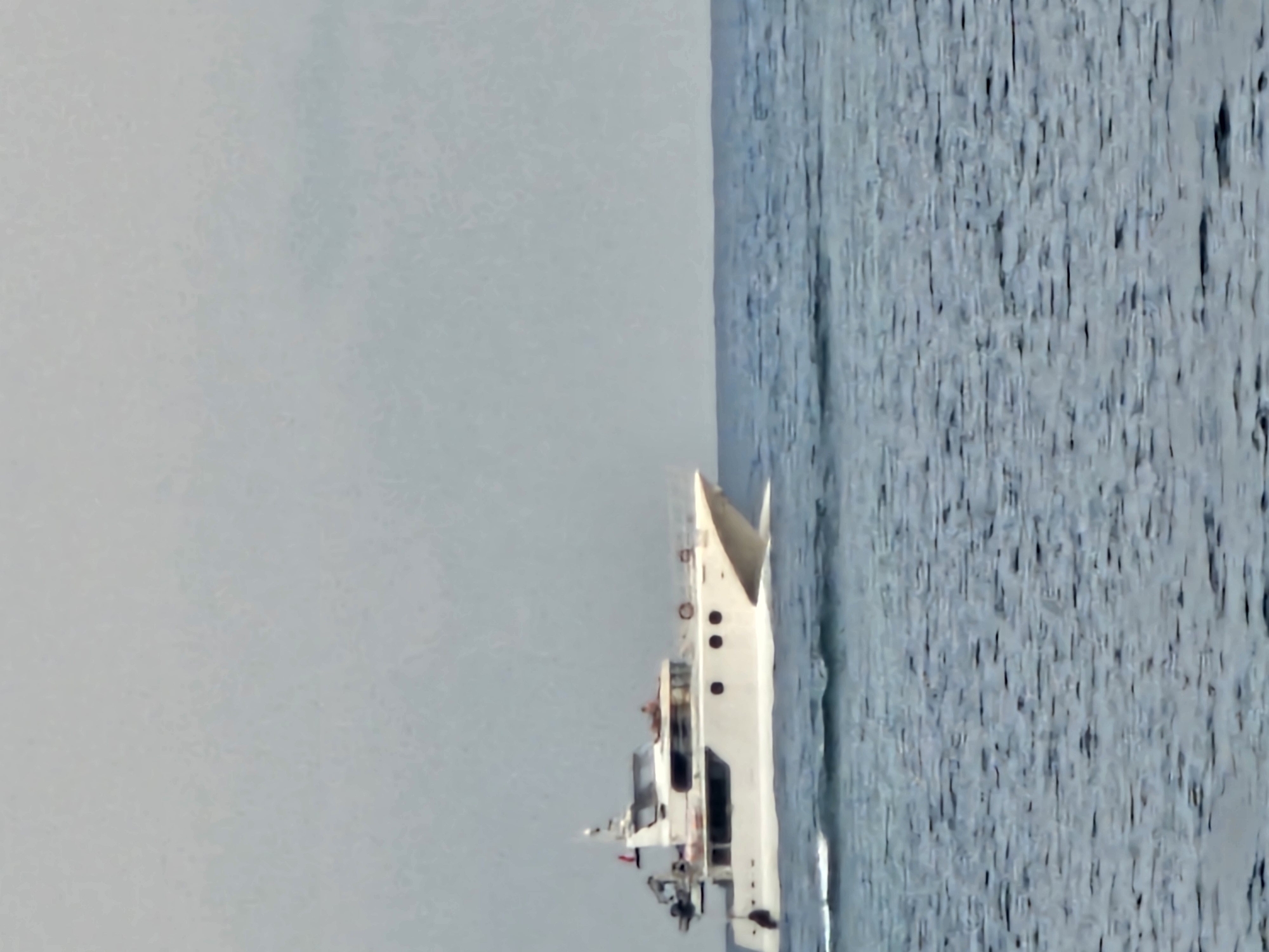 A white yacht cruises on the blue ocean under a cloudy sky. The yacht has multiple levels and appears to be moving from left to right. A person can be seen on the upper deck of the yacht. The ocean has moderate waves. The background consists of a light gray, overcast sky with no visible landmass. The image does not depict any identifiable landmarks or features that would place it specifically in Denpasar, Indonesia.