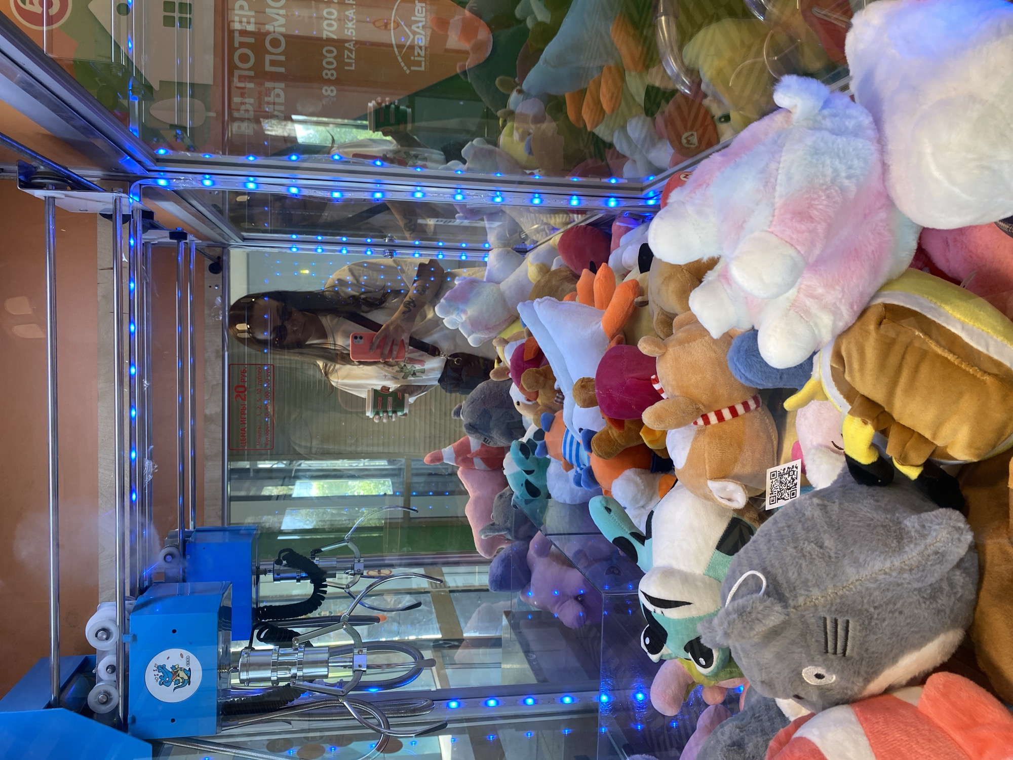 Here's a matter-of-fact description of the image:
**Scene:** The image shows the interior of a claw crane machine filled with plush toys. There are various stuffed animals visible, including a shark, a unicorn, and other generic plush figures. The machine is illuminated with blue LED lights along the edges.
**People:** A person with long brown hair is reflected in the glass of the crane machine, holding a pink phone and taking a picture.
**Objects:** Besides the plush toys and LED lights, there is the blue-colored claw mechanism of the machine. The crane's operational components (cables, pulleys, etc.) are visible. There is text in Russian on the glass.
**Environment:** The scene appears to be indoors in Votkinsk, Russia, evidenced by the Russian text on the machine.