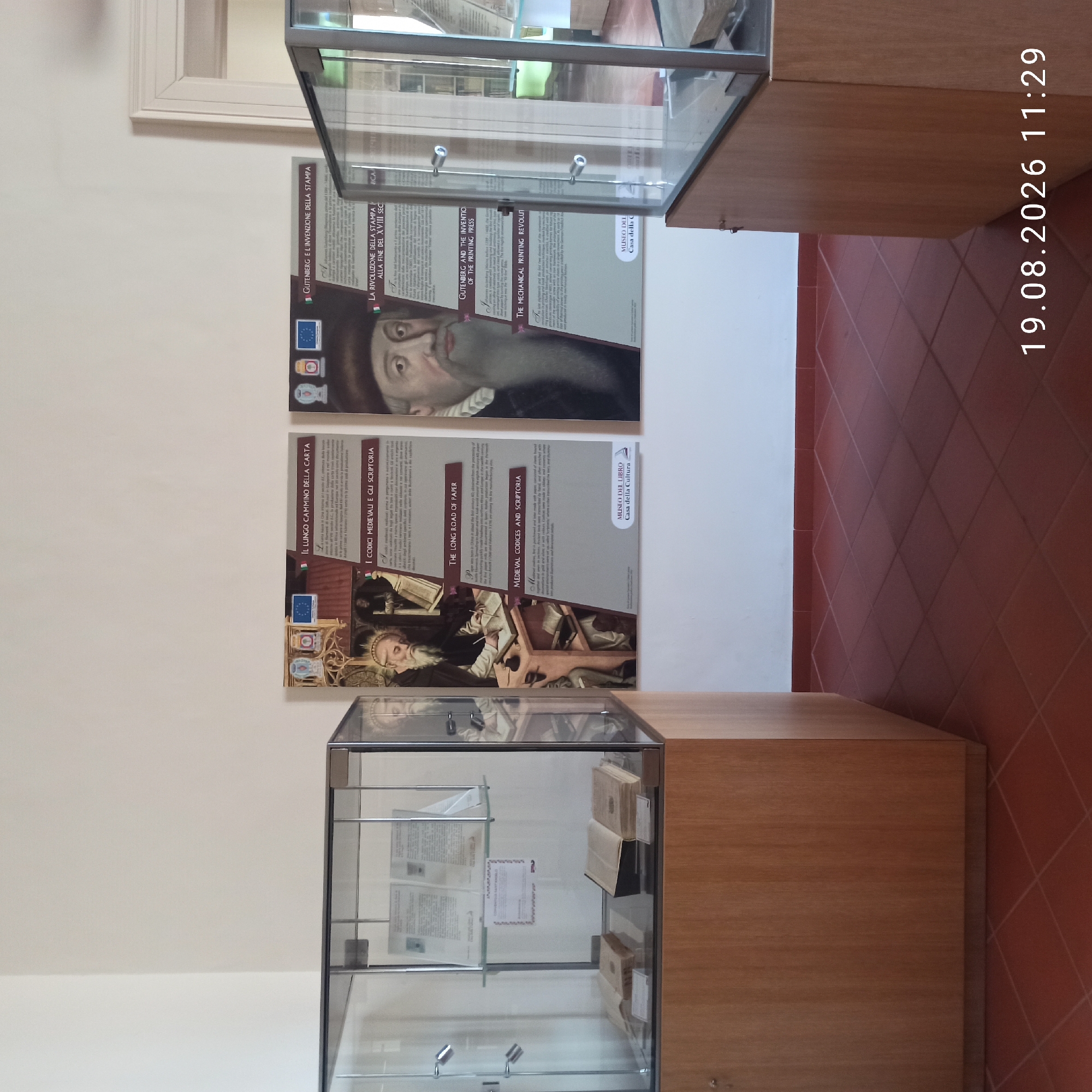 The image depicts an interior scene, likely a museum or exhibition space, featuring display cases filled with historical books and documents. The walls are painted a neutral color, and the floor is tiled in a terracotta diamond pattern. In the background, there are informational posters with text in Italian, discussing topics related to the history of paper and printing. One poster features a portrait of Johannes Gutenberg.
One of the display cases in the foreground contains several open books with aged pages, some with visible script or print. Another case holds what appear to be documents or leaflets displayed upright on shelves. A third display case, partially visible on the right side of the image, also appears to house books. The overall impression is of a well-maintained exhibition focusing on historical artifacts related to writing and printing.
The image was taken on August 19, 2026, at 11:29. While the image does not explicitly state the location within Ruvo di Puglia, Italy, the presence of Italian text on the posters suggests a connection to Italy. Such an exhibition is likely to be housed in a cultural institution within the city.