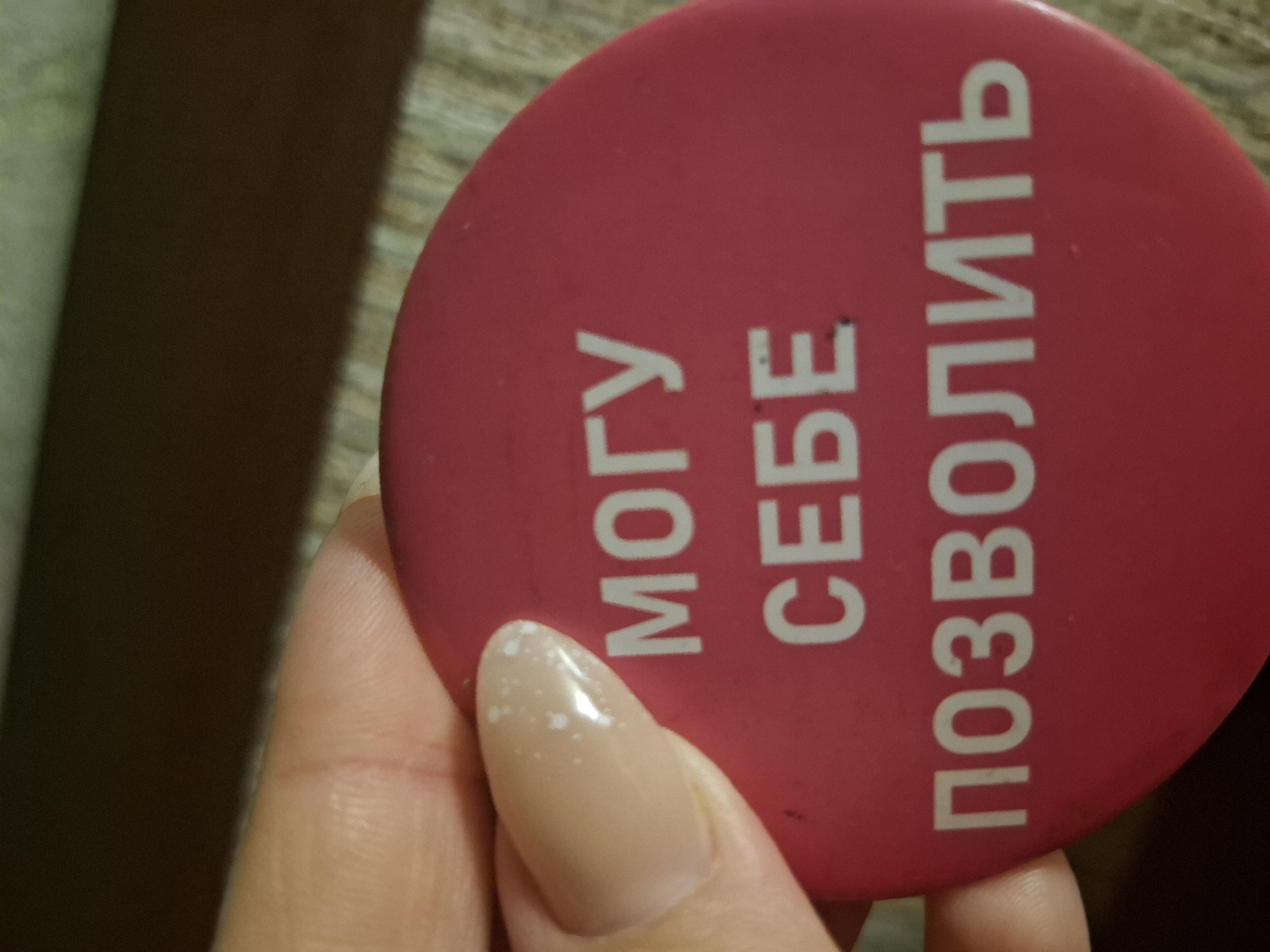 A close-up shot features a human hand holding a bright fuchsia-colored, circular object. The visible parts of the hand include a thumb and an index finger, both adorned with long, almond-shaped nails. These nails are painted in a light beige or nude polish, with the thumbnail showing several small, irregular white specks, possibly indicating chipped polish or a decorative element.
The round object, which appears to be a pin, badge, or coaster, is angled in the hand. It bears prominent white Cyrillic text arranged vertically along its surface. The text reads "МОГУ СЕБЕ ПОЗВОЛИТЬ," which directly translates to "I CAN AFFORD IT" or "I CAN ALLOW MYSELF." The lettering is sharp and clear against the vibrant pink background of the object.
The background is softly blurred and out of focus, displaying muted tones of dark brown and lighter beige, suggesting an indoor setting with possibly wooden furniture or textured surfaces. The lighting in the scene appears to be dim or artificial, contributing to the intimate, close-up nature of the photograph. The presence of Cyrillic text strongly indicates a connection to a Russian-speaking region, aligning with the specified location of Markovskiy, Russia. The image captures a simple, personal moment of holding an object with an affirmative message.